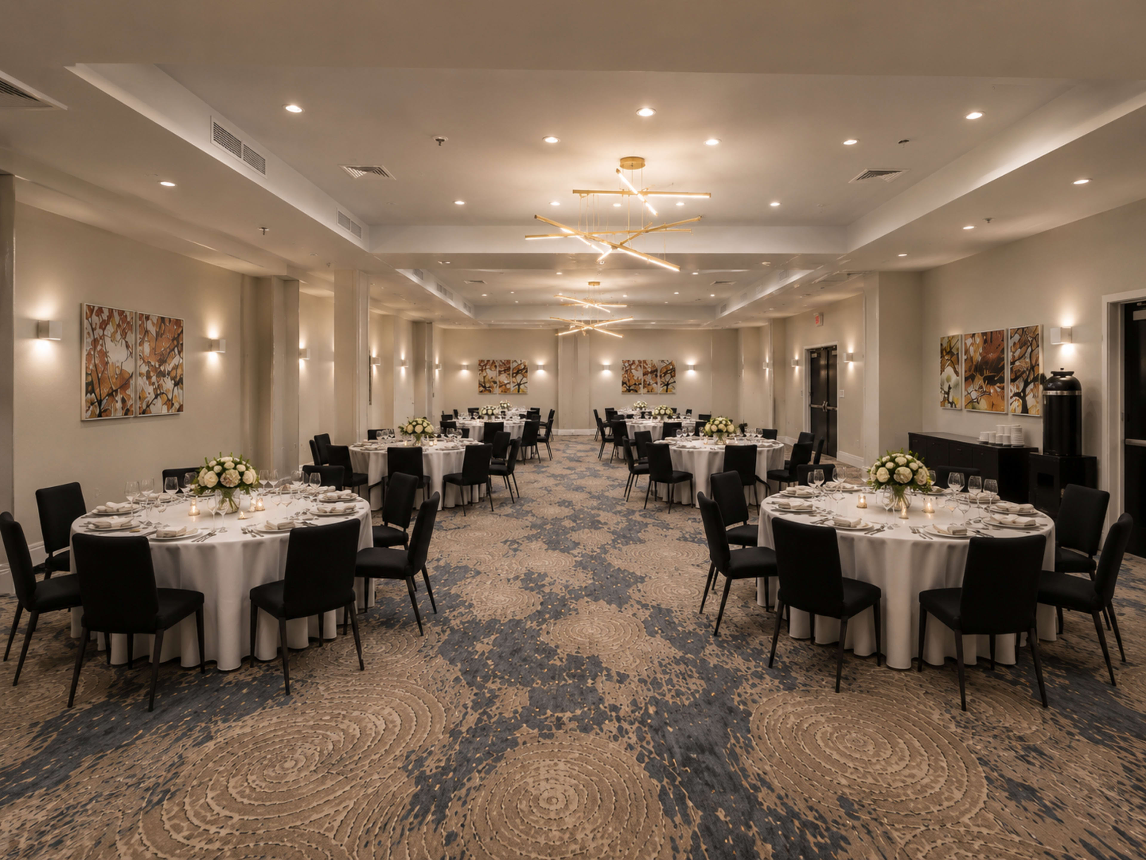 The image shows a banquet hall set up with round tables adorned with white tablecloths and floral centerpieces, illuminated by modern light fixtures.