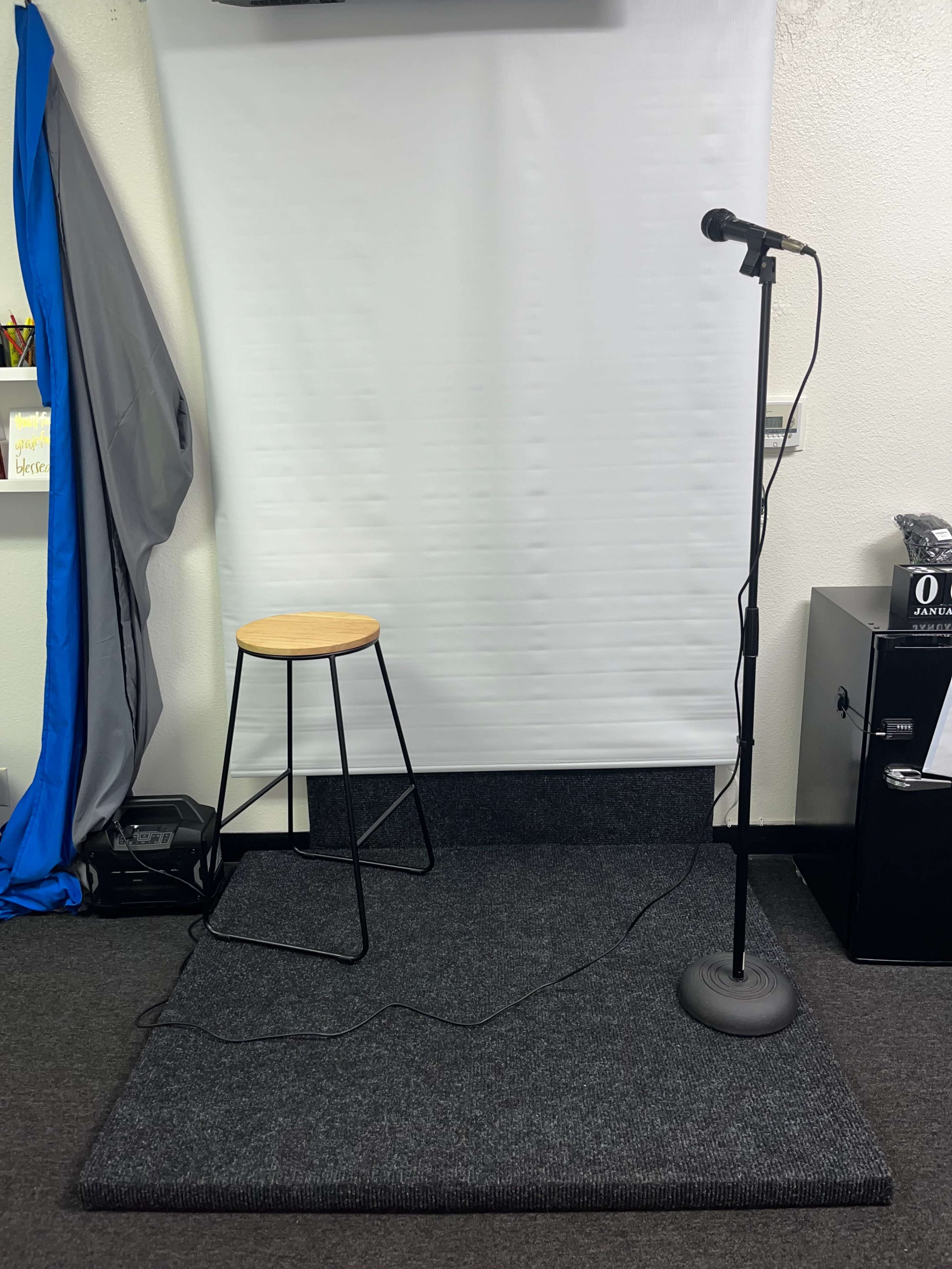 A small stage setup featuring a microphone on a stand next to a stool in front of a white backdrop.