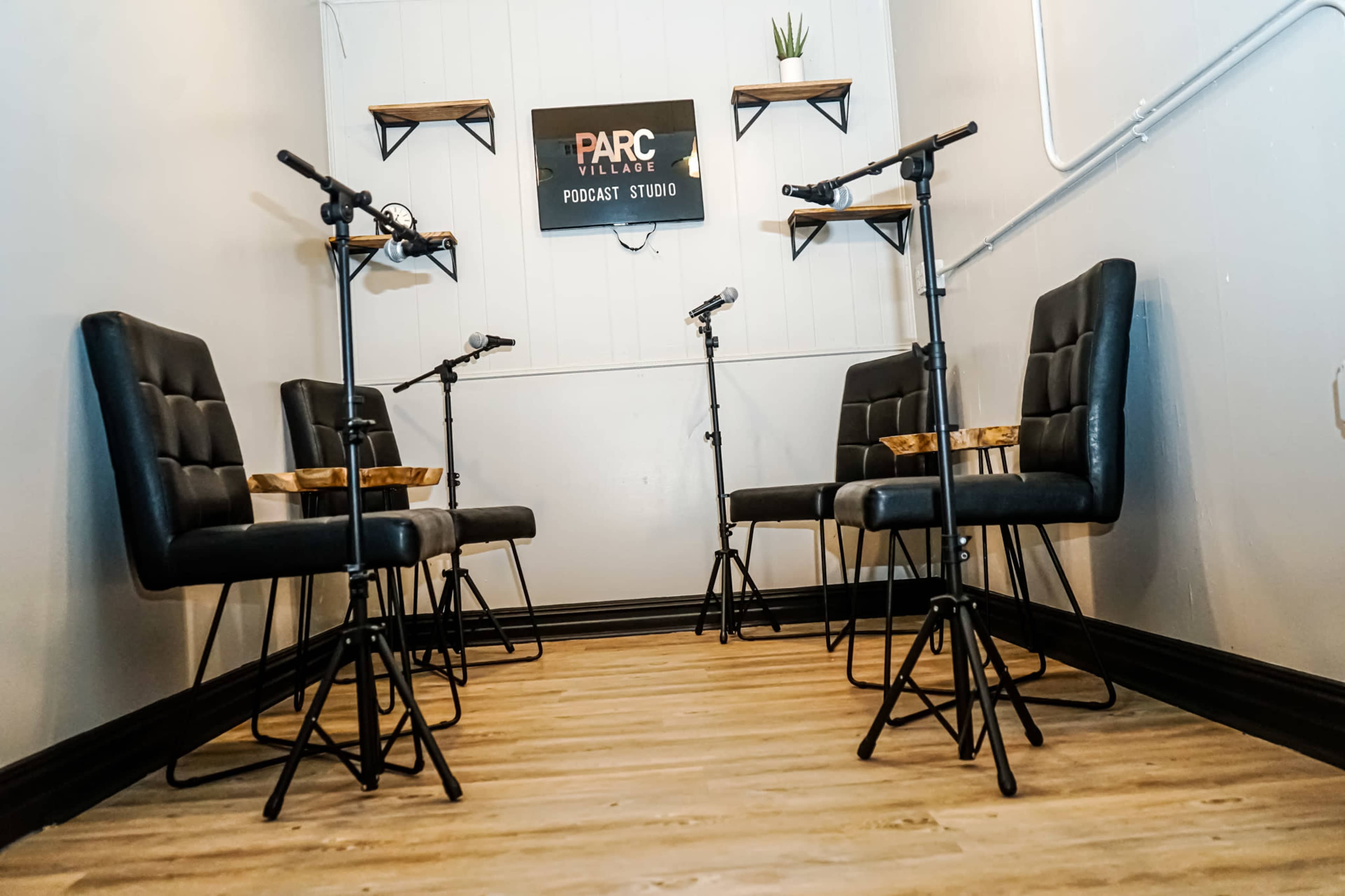 The image shows a small podcast studio featuring four black chairs arranged around a central space, with microphones mounted on stands and shelves displaying decor on the walls.