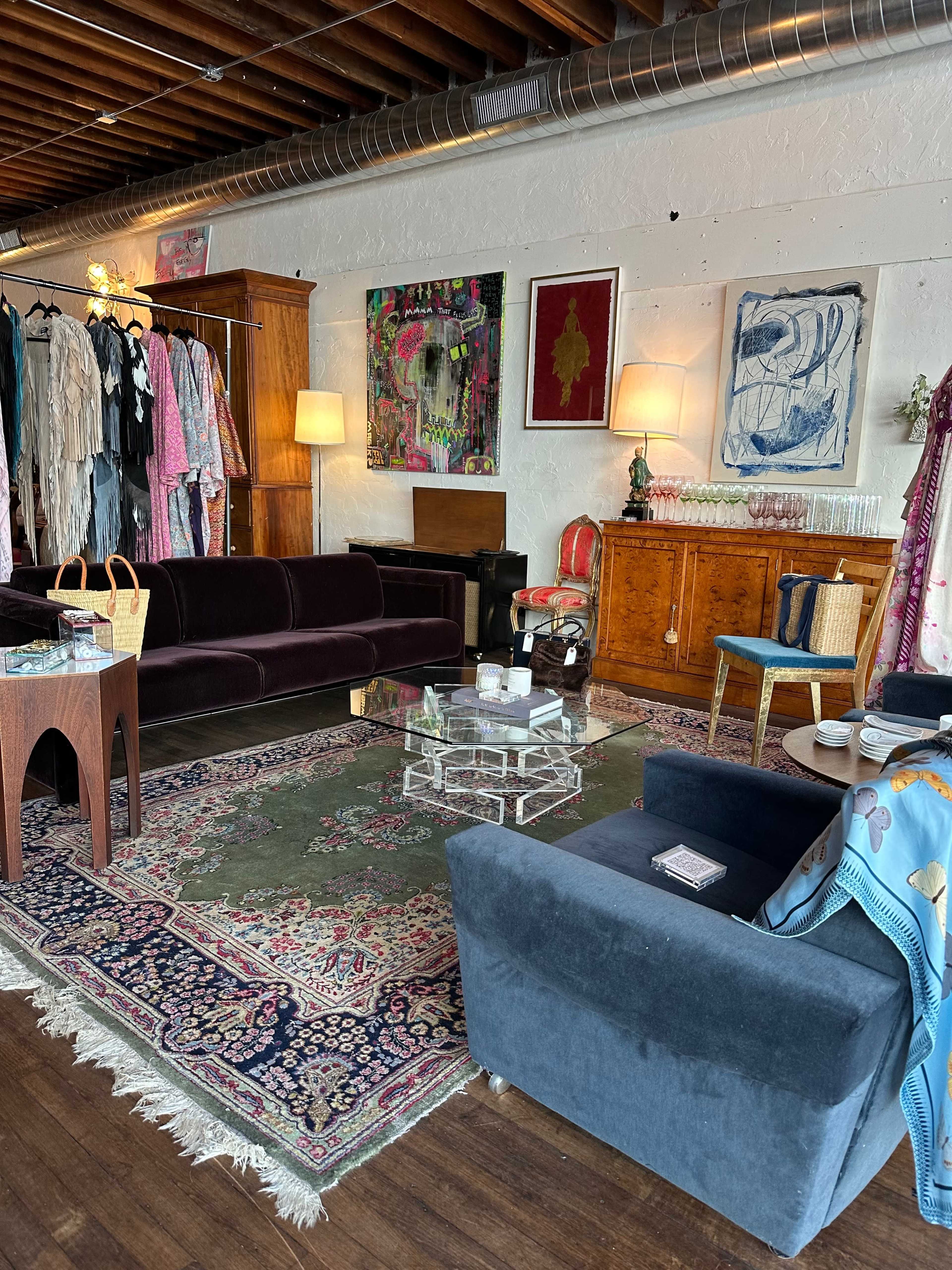A stylish boutique interior featuring a mix of clothing racks, artwork on the walls, a plush sofa, and a glass coffee table set on a patterned rug.