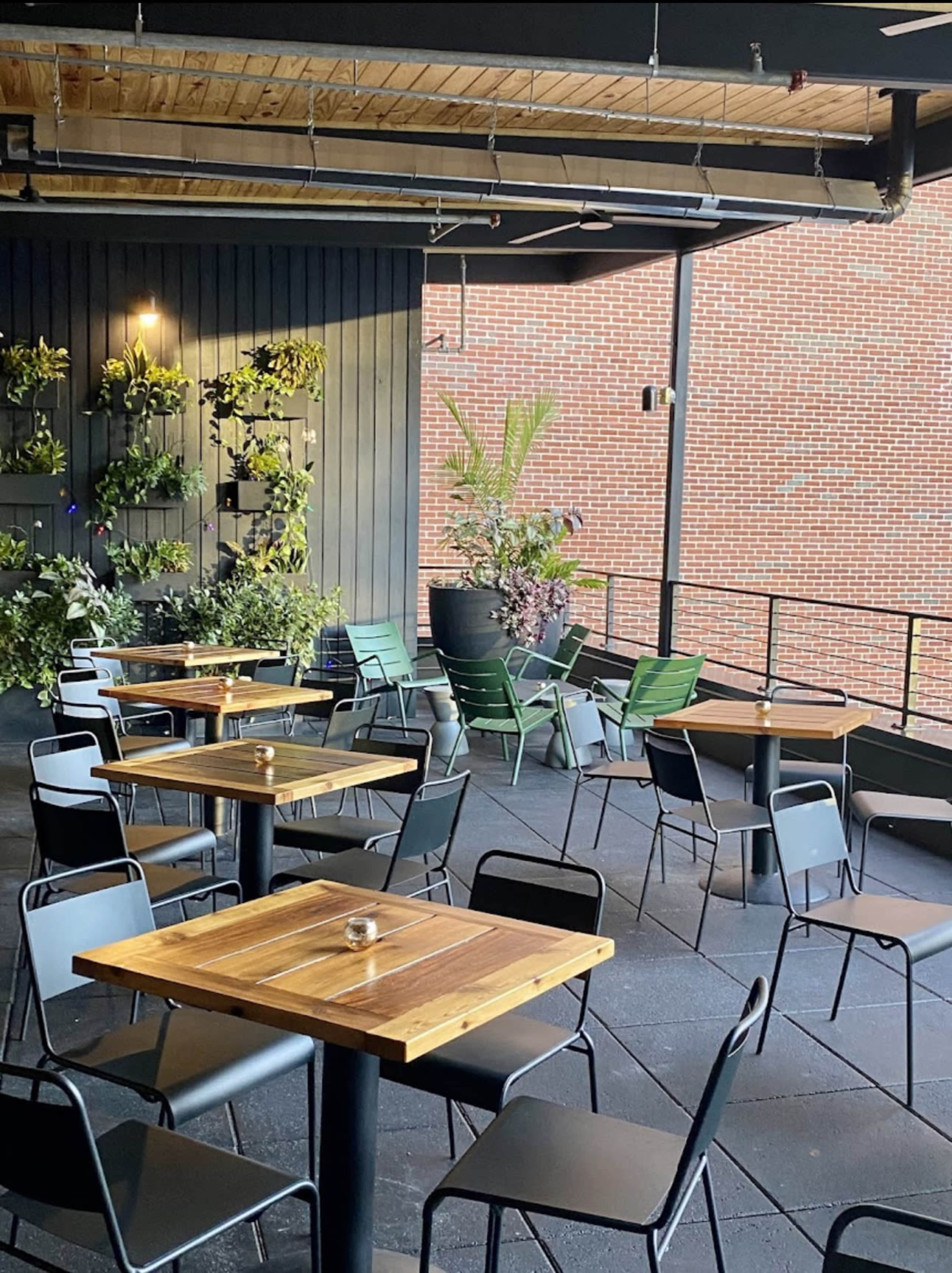 A modern outdoor seating area features wooden tables and black metal chairs, surrounded by greenery and brick walls.