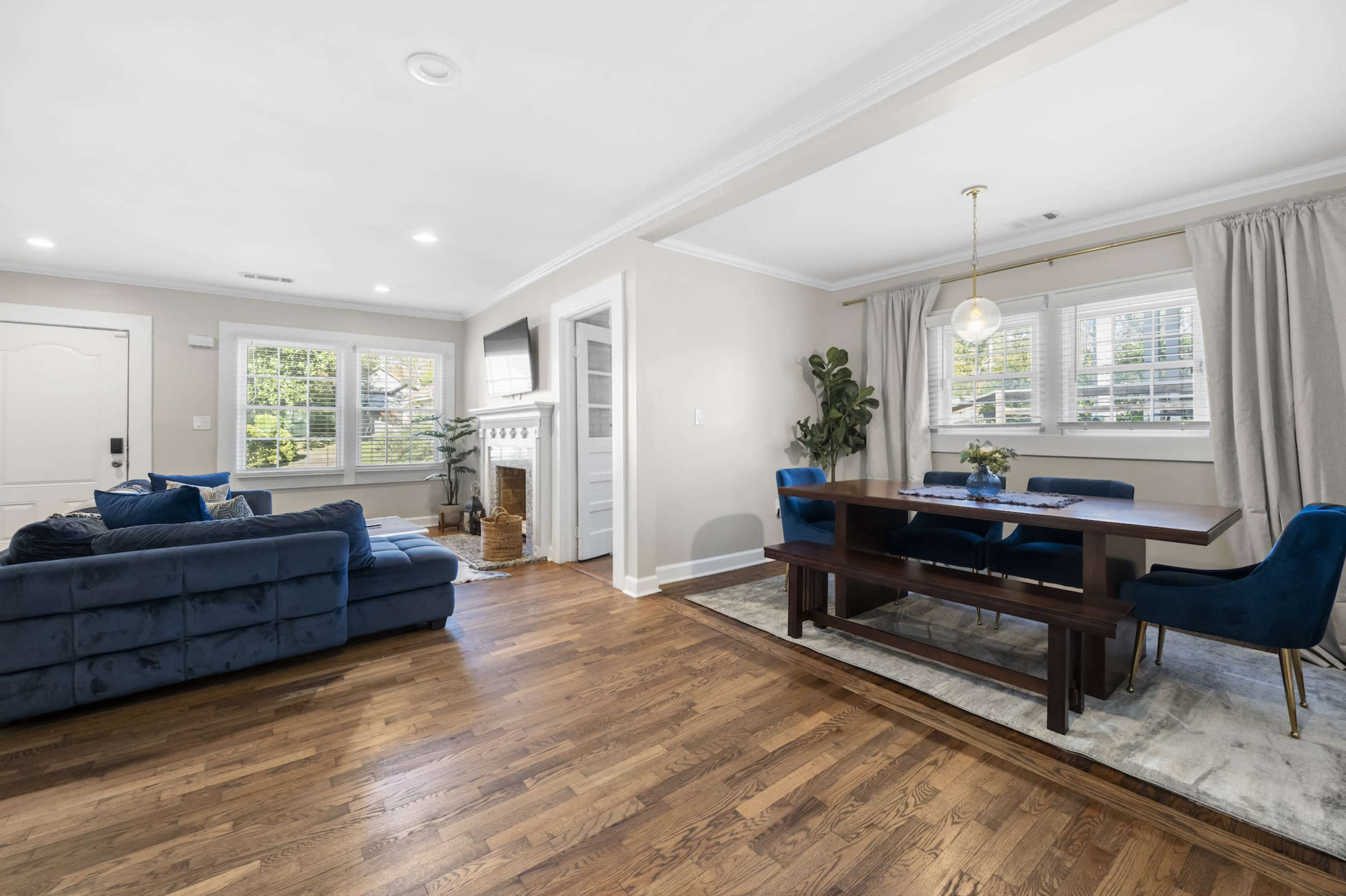 A living room and dining area feature a blue sectional sofa, a wooden dining table, and large windows with white curtains.