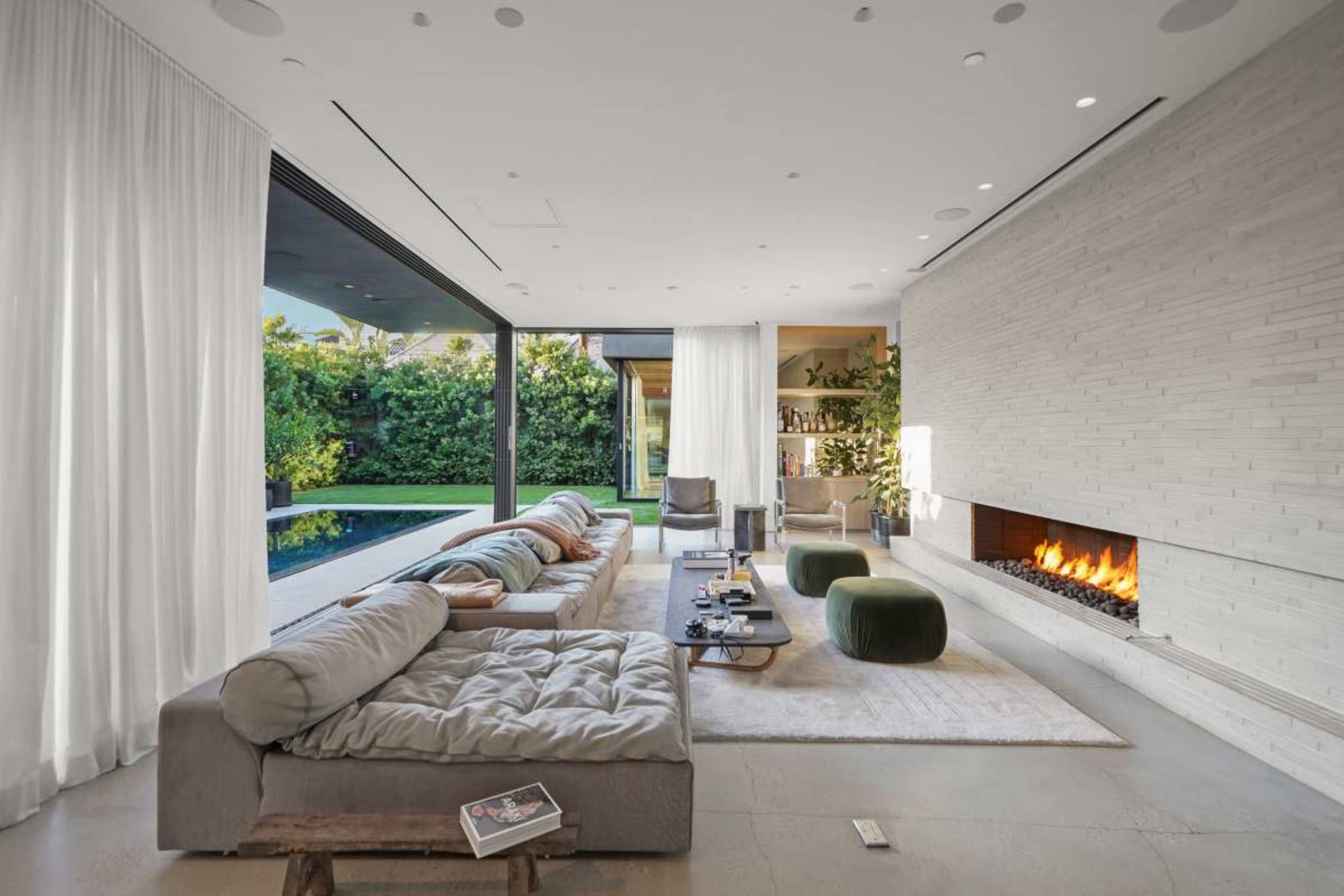 The image shows a modern living room with a sectional sofa, a fireplace, large glass windows, and a view of a garden and pool.