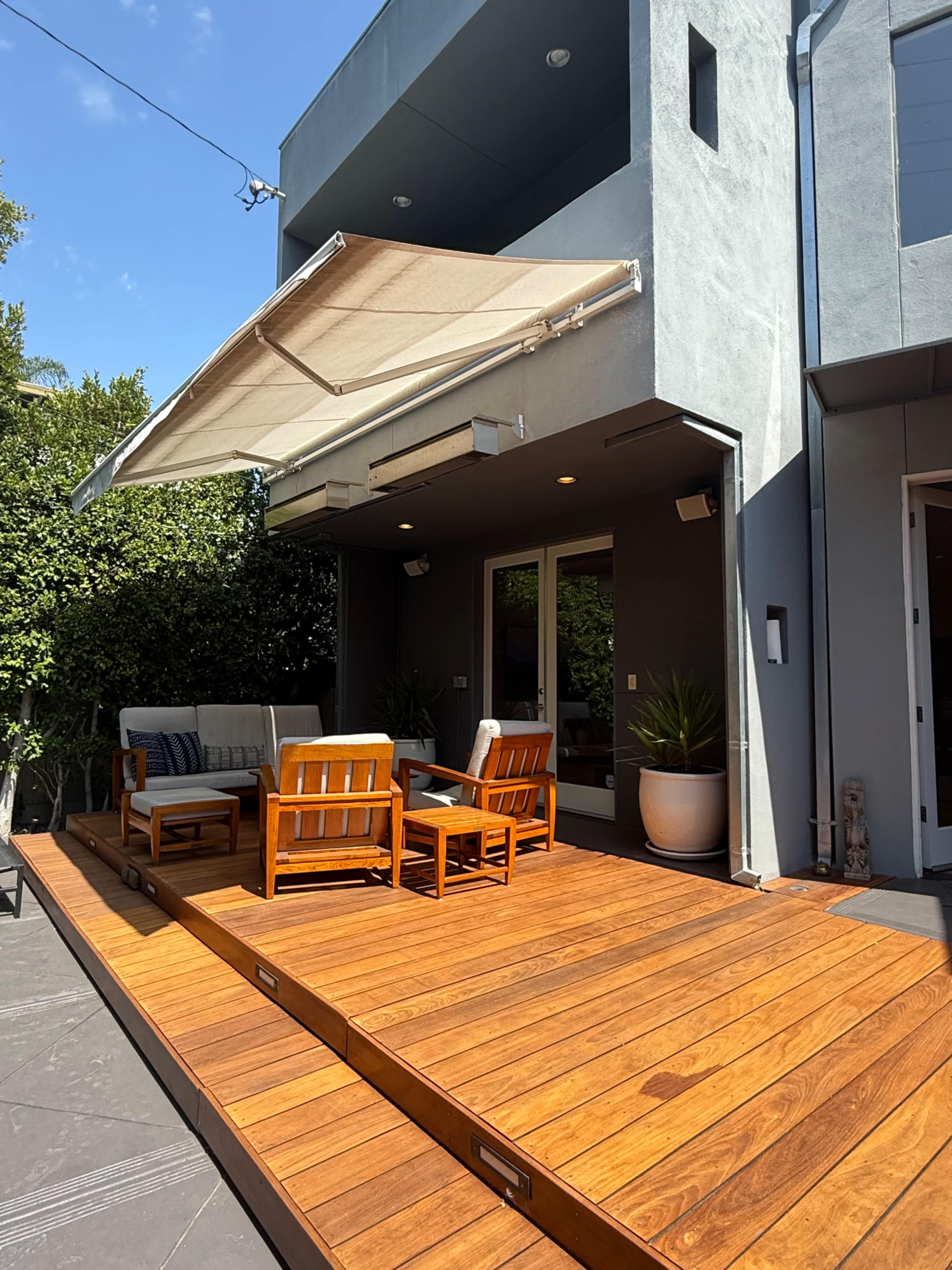 The image features a modern wooden deck with two lounge chairs and a canopy extending over a seating area next to a house surrounded by green foliage.