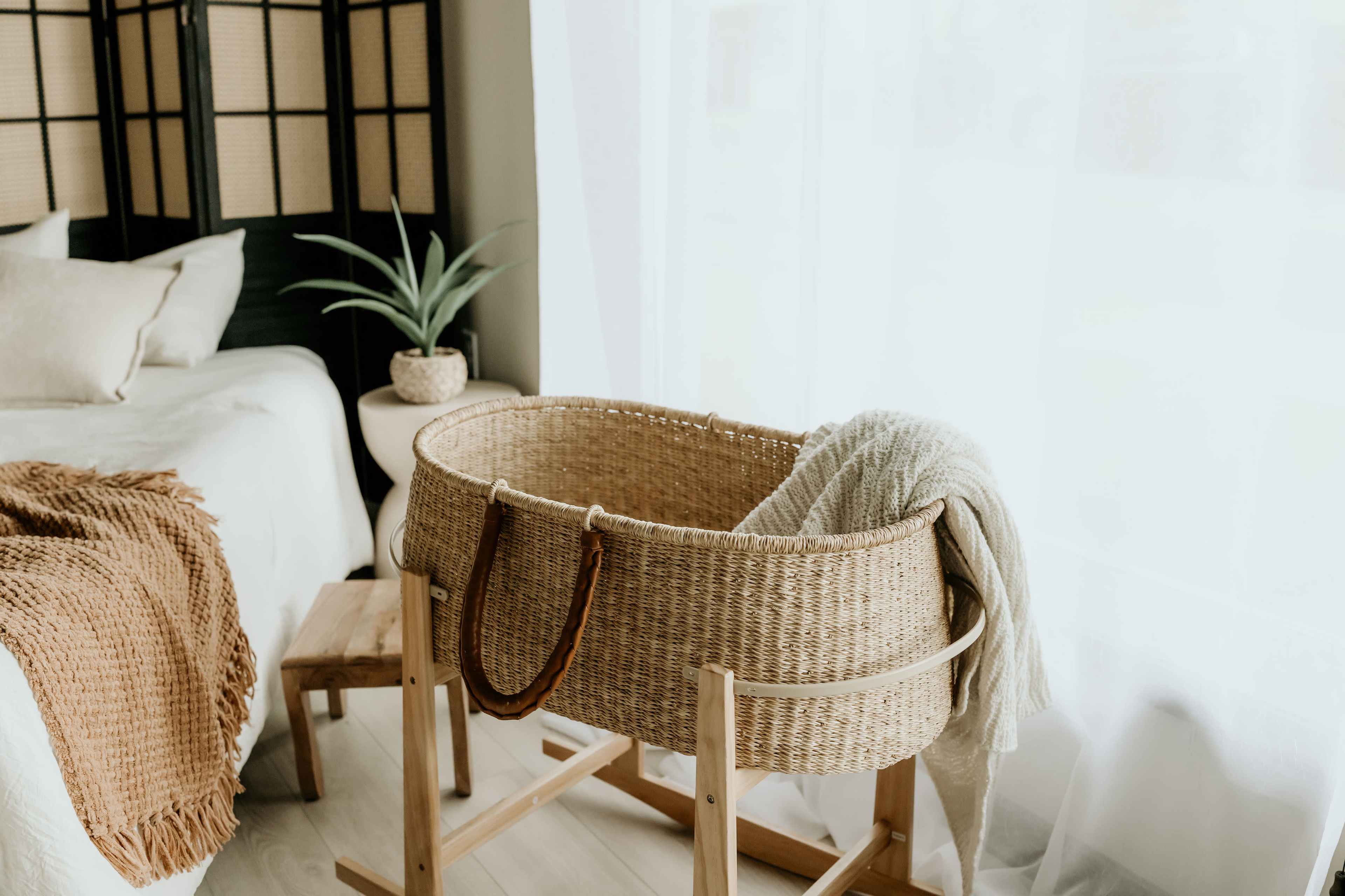 A woven bassinet with a blanket draped over it is positioned next to a bed in a softly lit room.