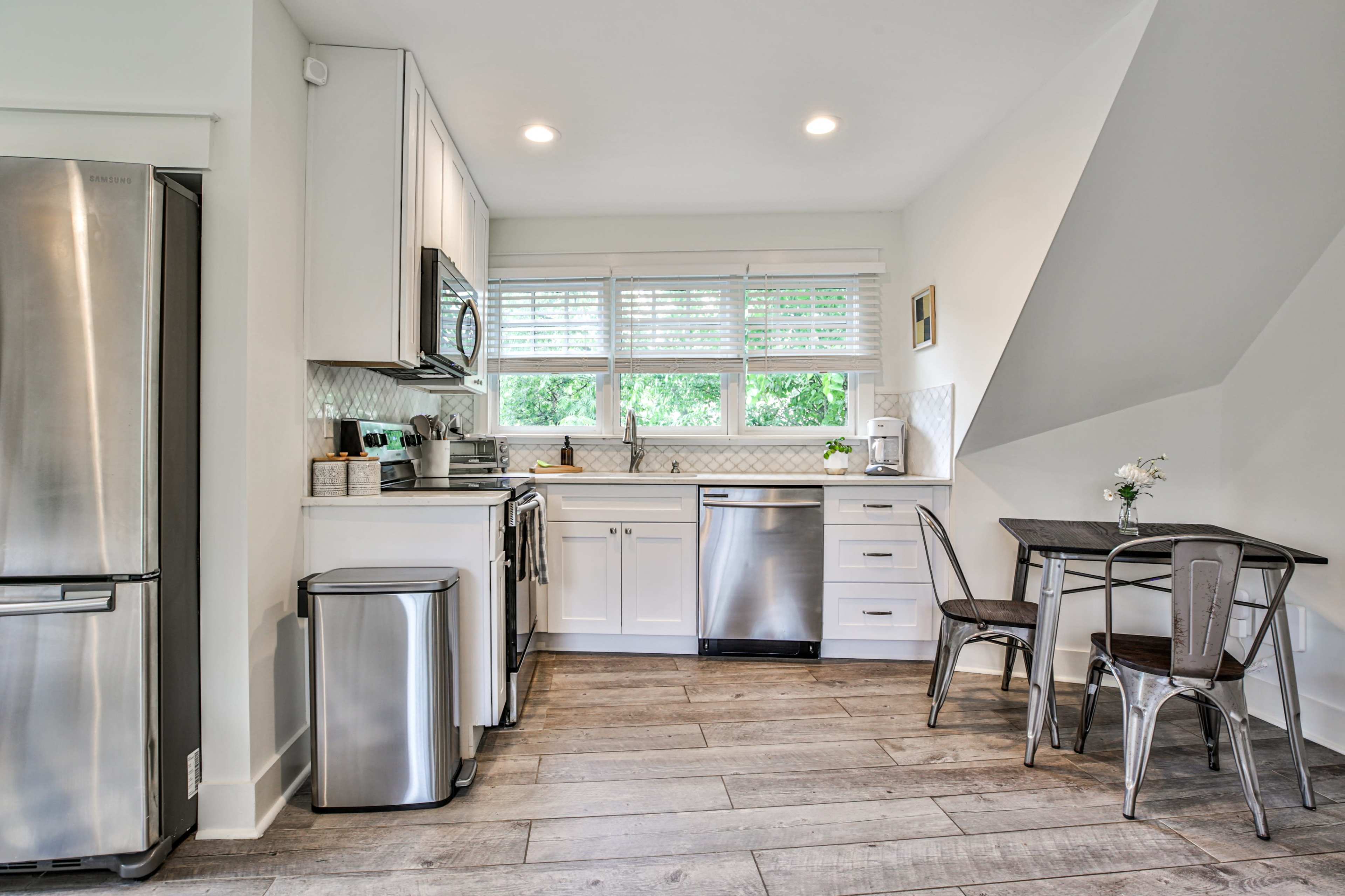 A modern kitchen featuring stainless steel appliances, white cabinetry, and a small dining table with metal chairs under a window.