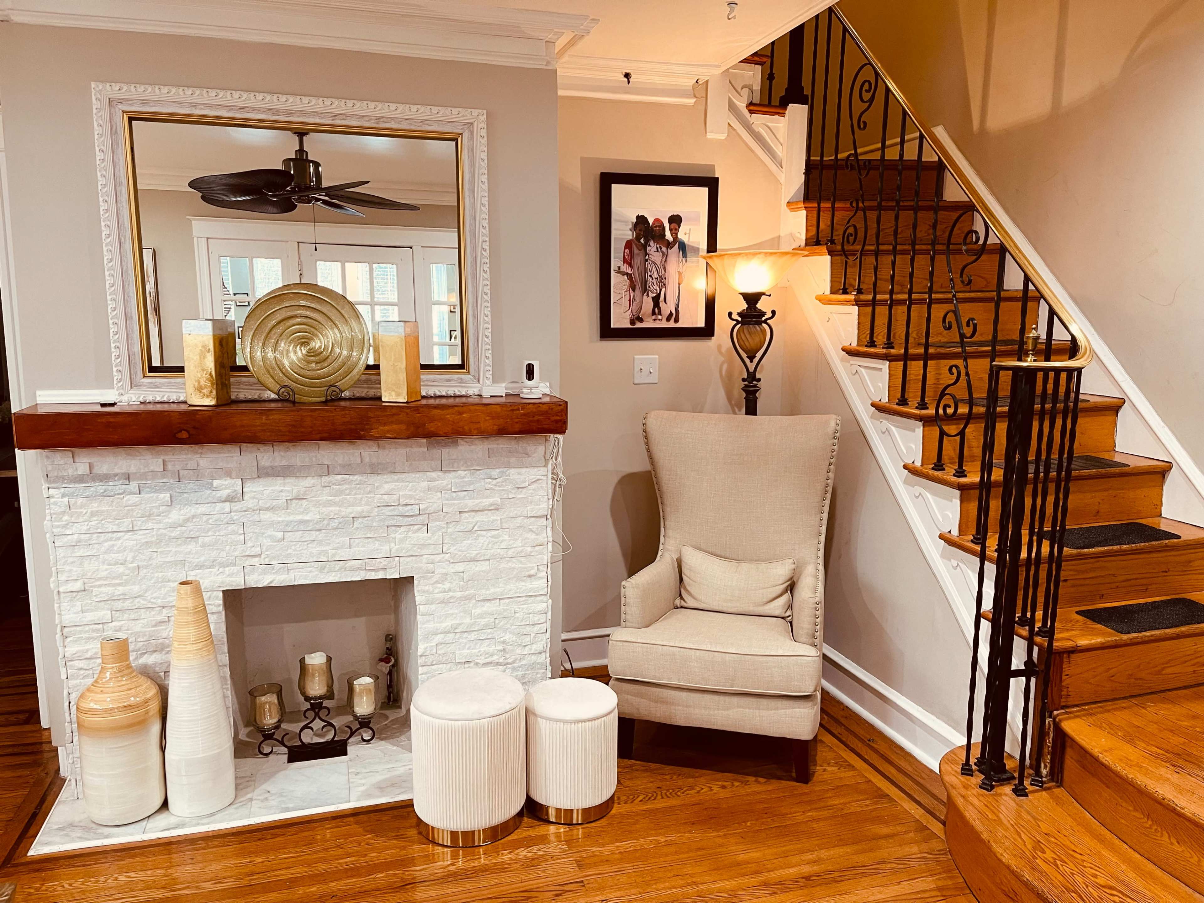 A living room features a light-colored stone fireplace with decorative vases and two stools beside a beige armchair, facing a staircase with wooden steps and a black railing.