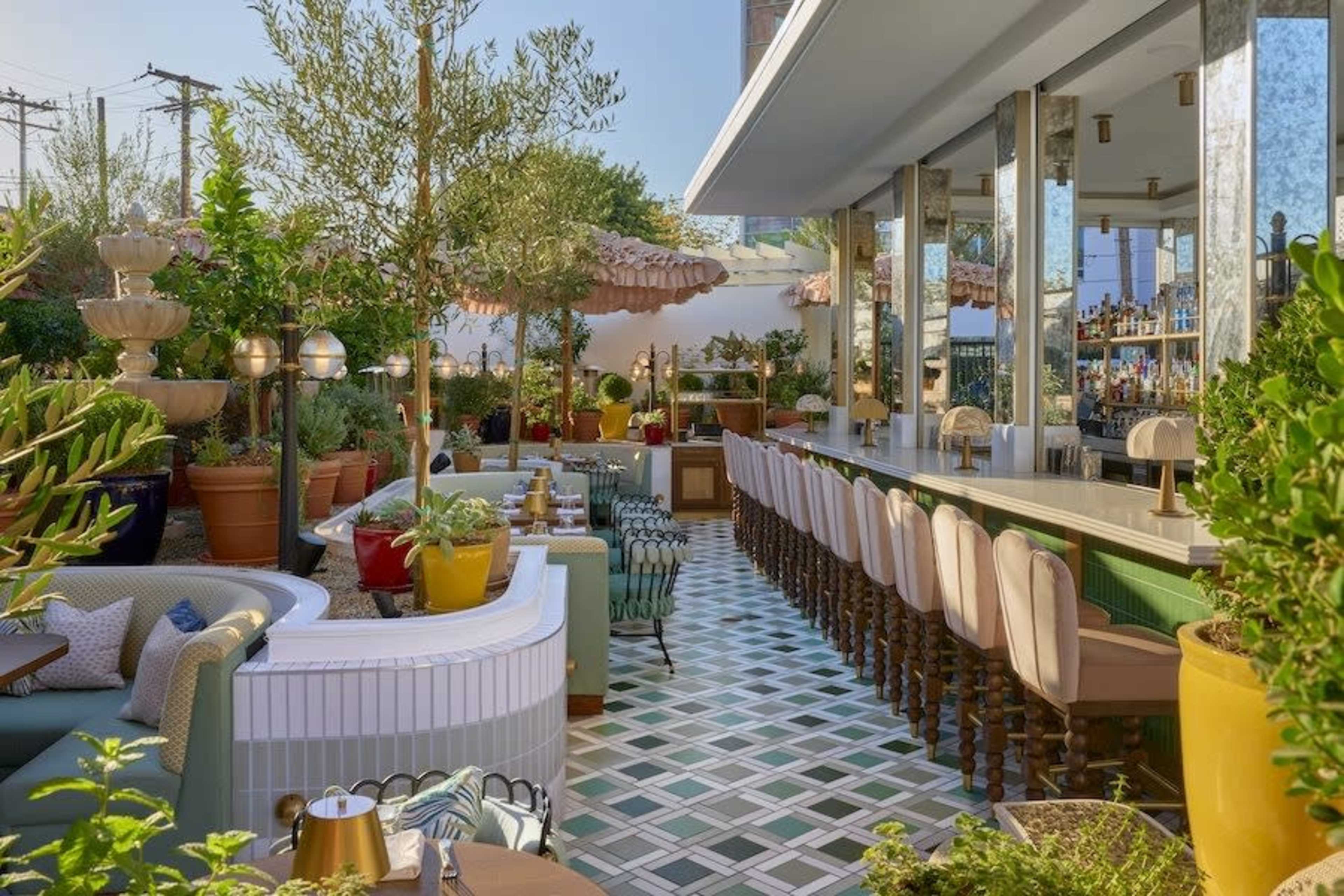 The image shows a stylish outdoor restaurant patio with a bar, surrounded by greenery and decorative plants.