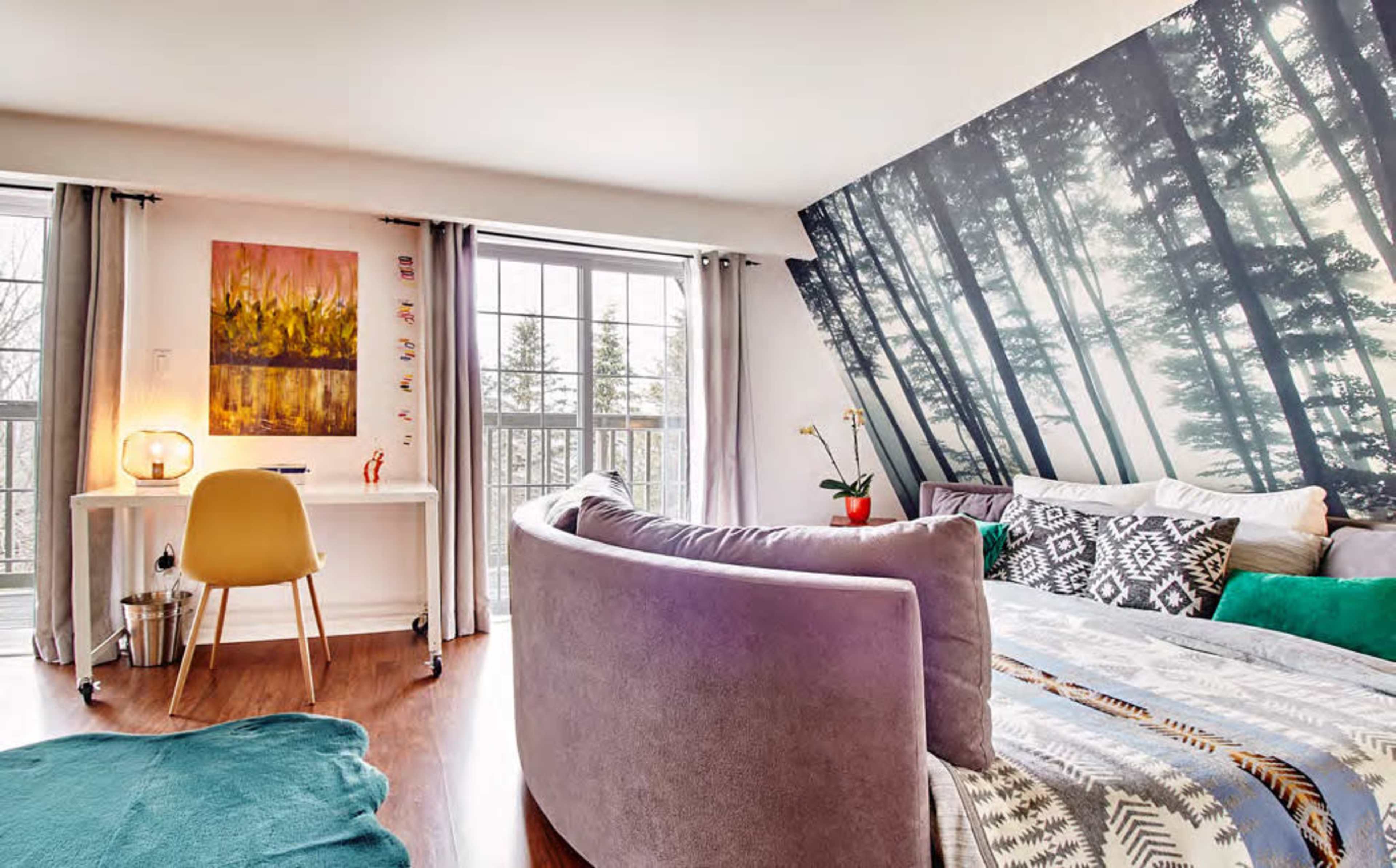 A modern bedroom features a curved sofa, a wooden desk with a lamp, and a large forest-themed mural on one wall.