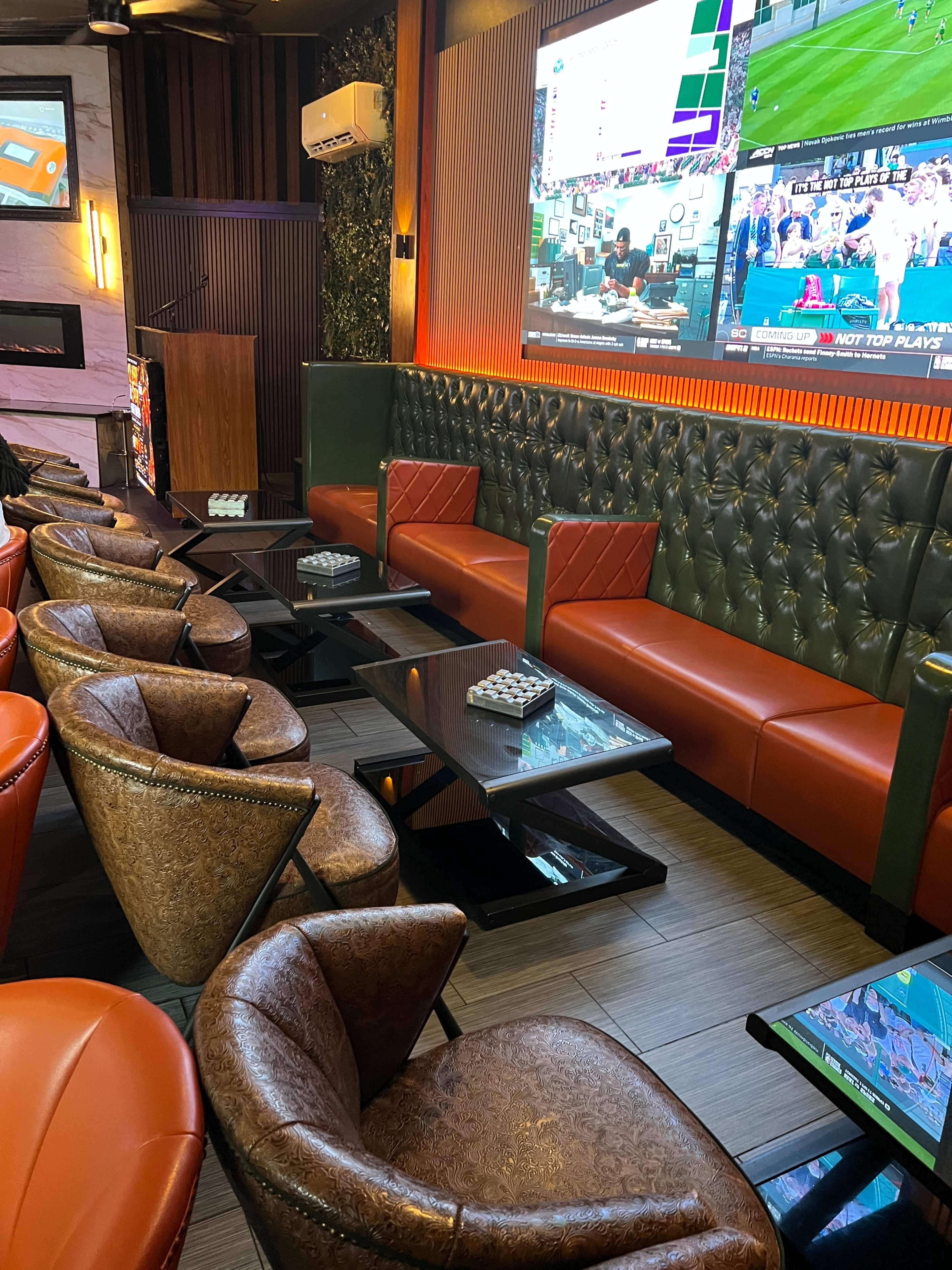 The image shows a modern lounge area with leather seating and multiple screens displaying sports events.