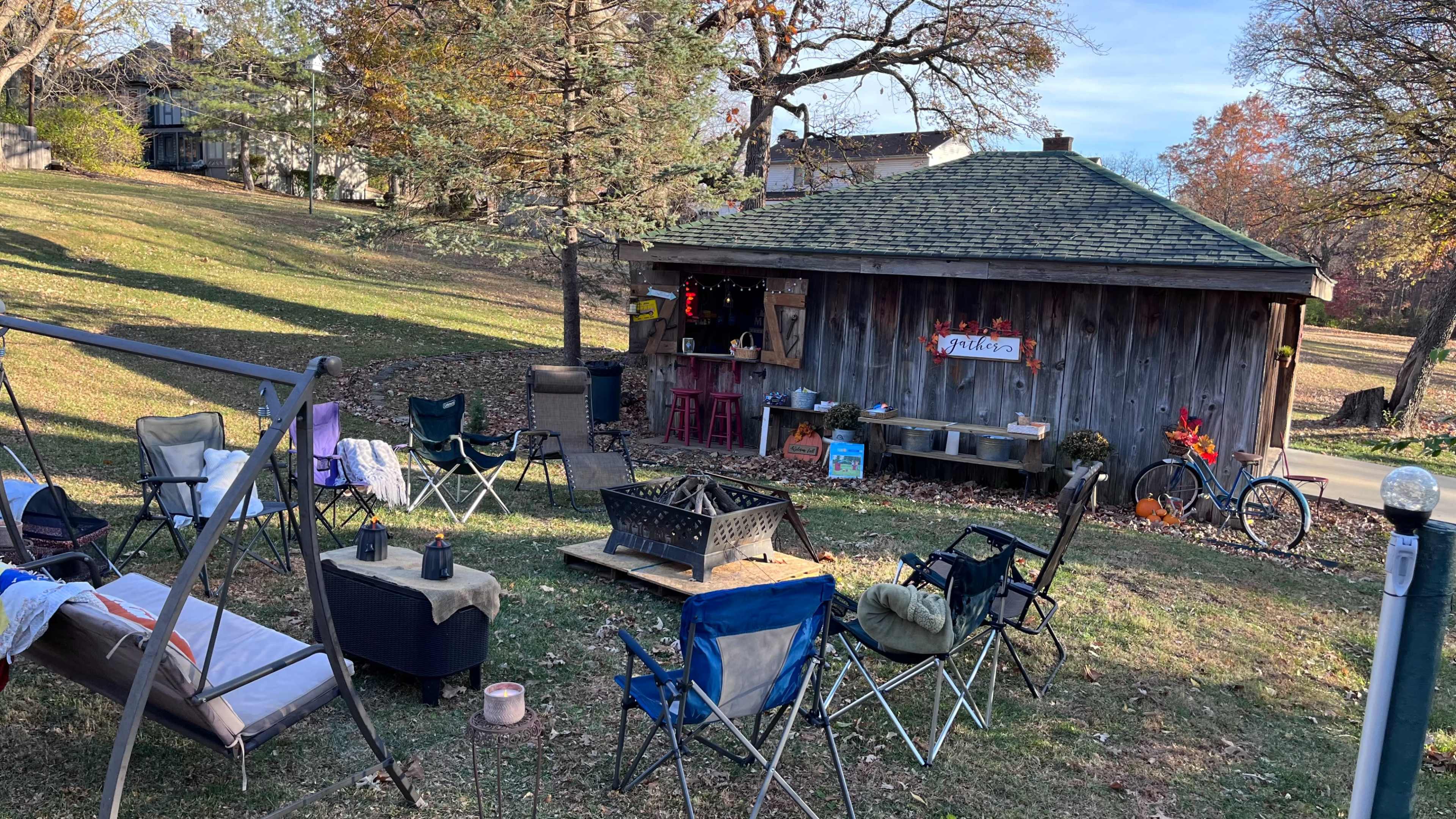 The image shows a backyard setup with several folding chairs arranged around a fire pit, adjacent to a rustic shed decorated for a gathering.