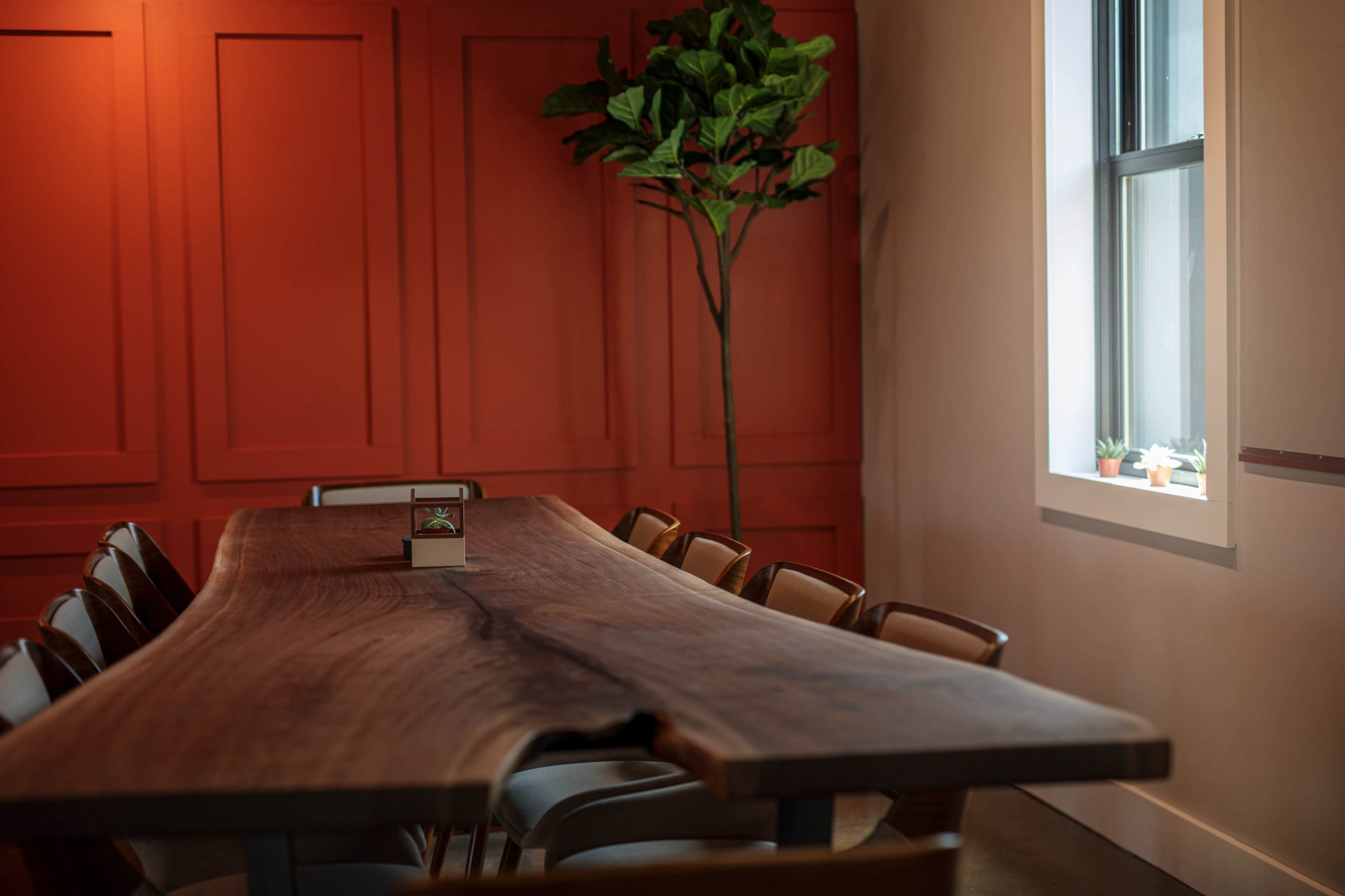 A large wooden table with several chairs sits in a meeting room featuring orange paneled walls and a small tree near the window.