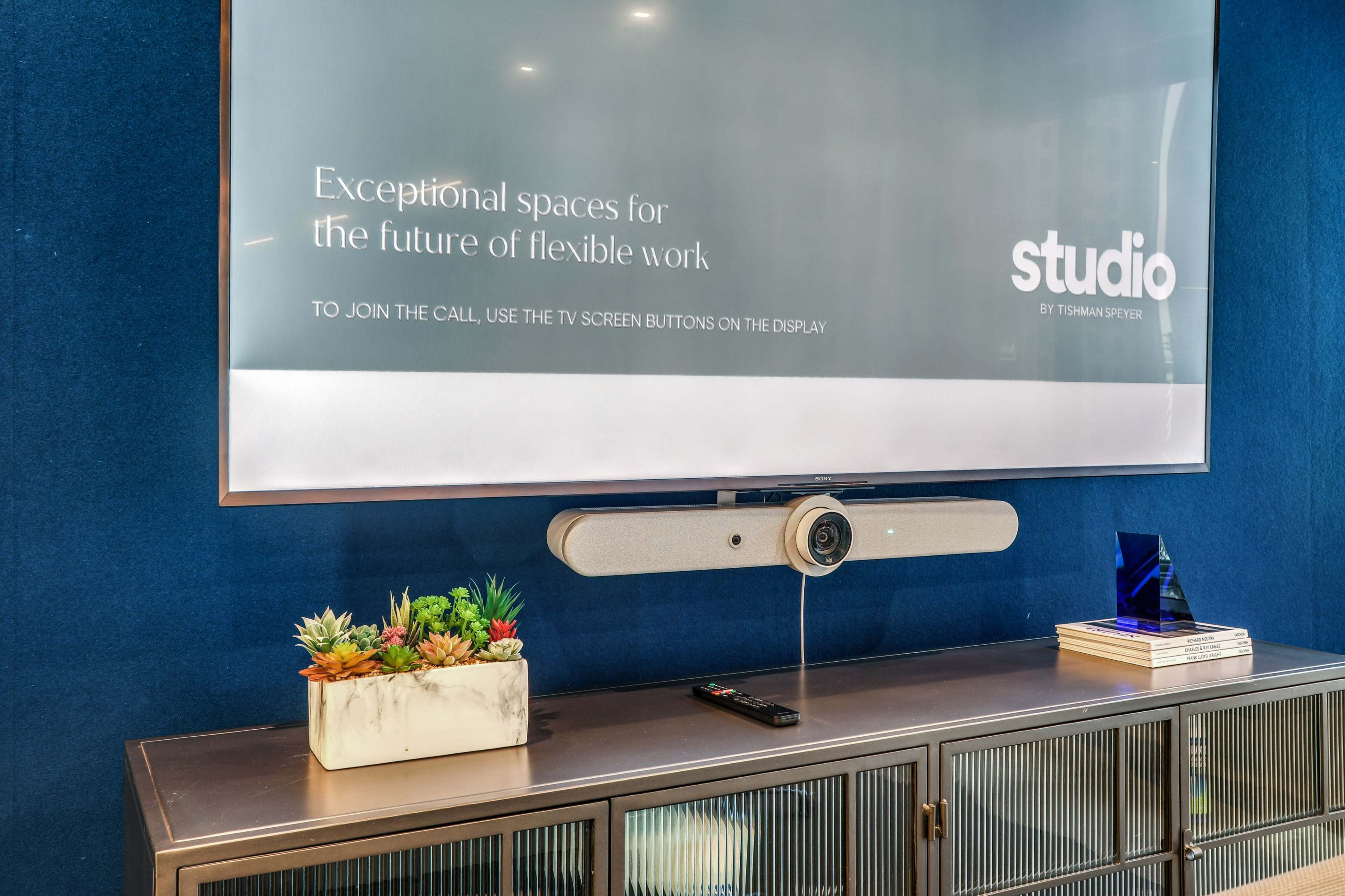 A modern conference room features a large screen displaying information about flexible workspaces, with a video conference system and a decorative plant on a cabinet below.