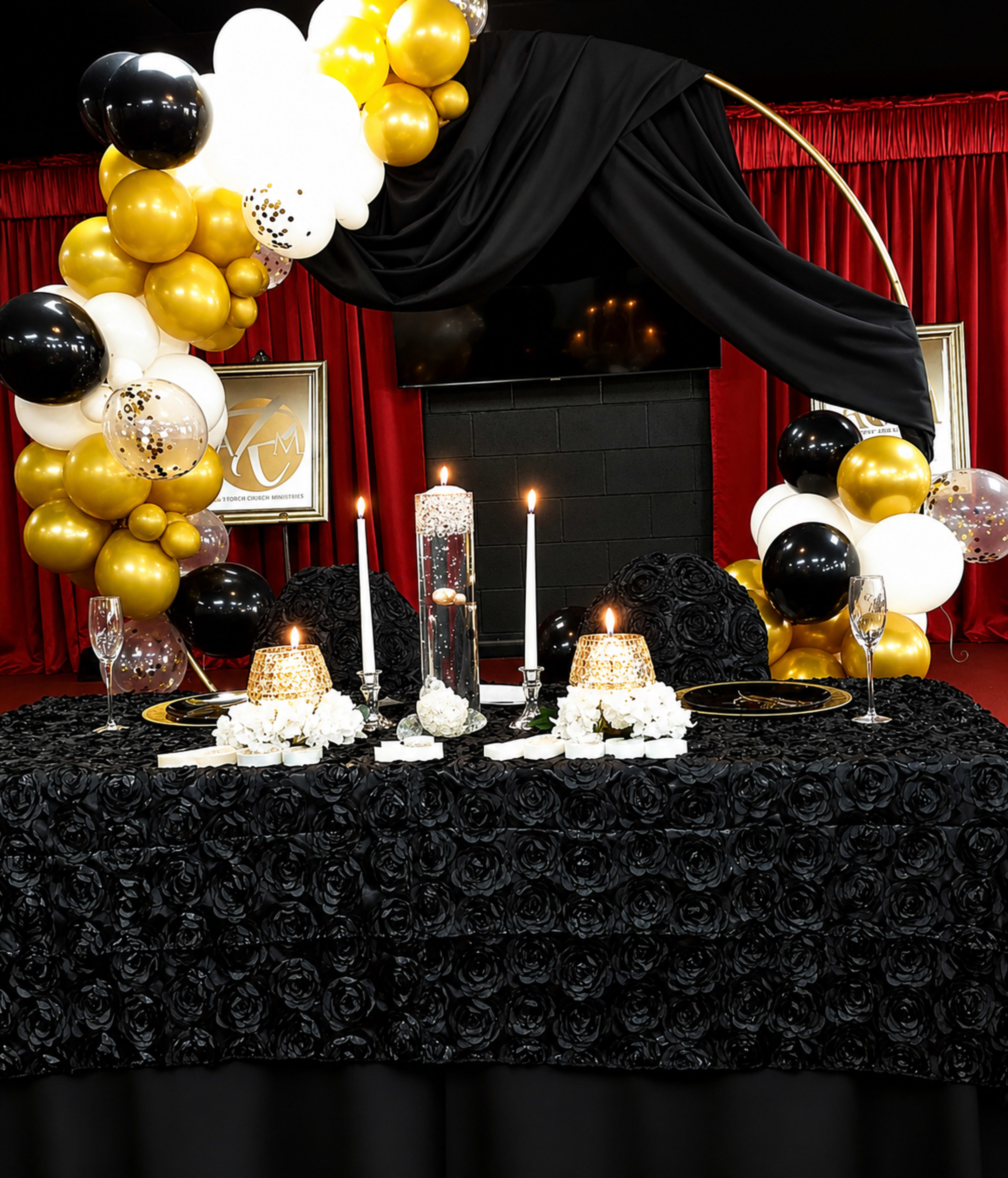 The image shows an elegantly arranged dining table decorated with a black rose-patterned tablecloth, gold and black balloons, lit candles, and floral accents against a red backdrop.