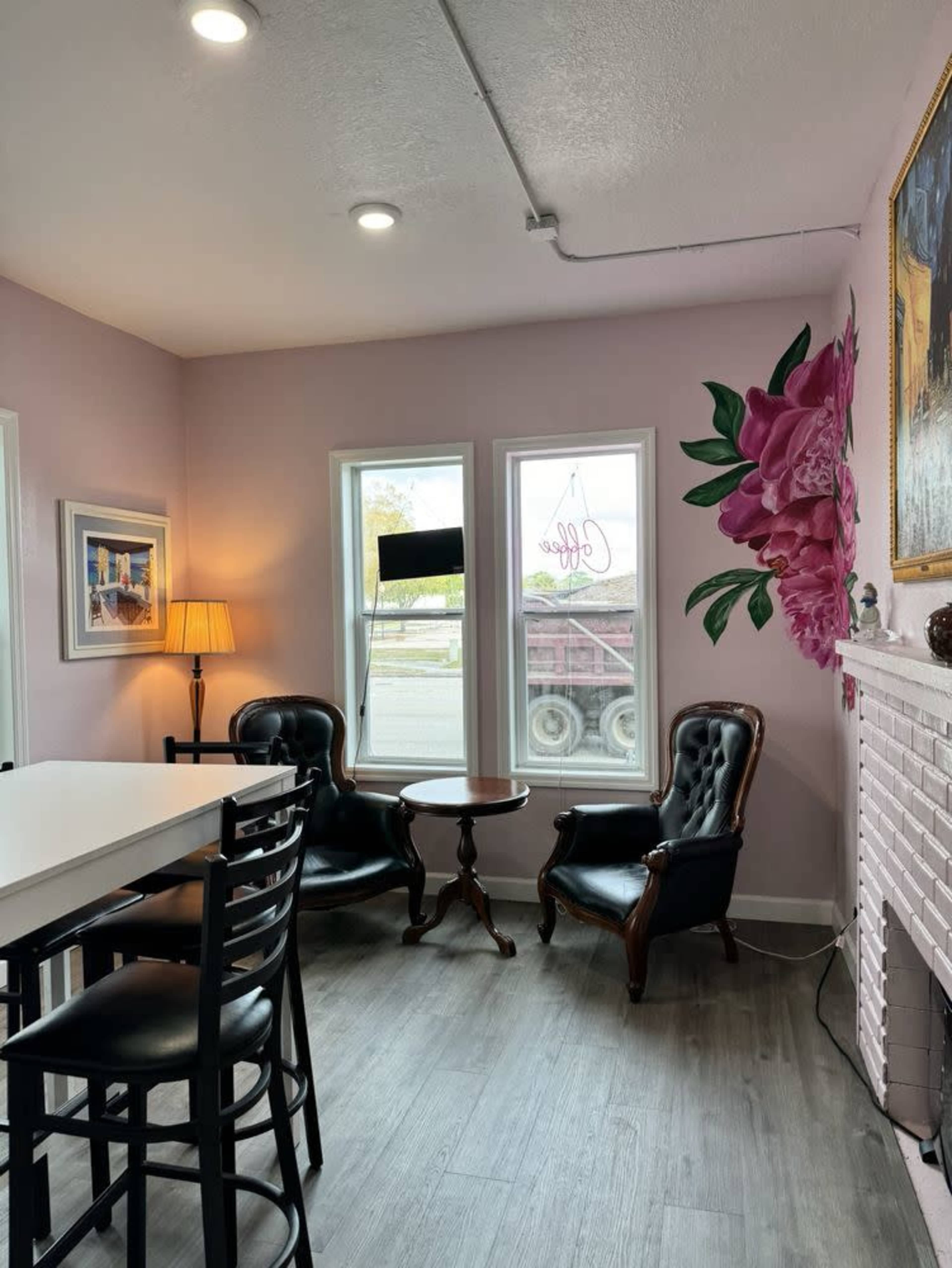 The image shows a small café interior with two black leather chairs, a round table, and a light-colored wall decorated with a large pink flower mural.