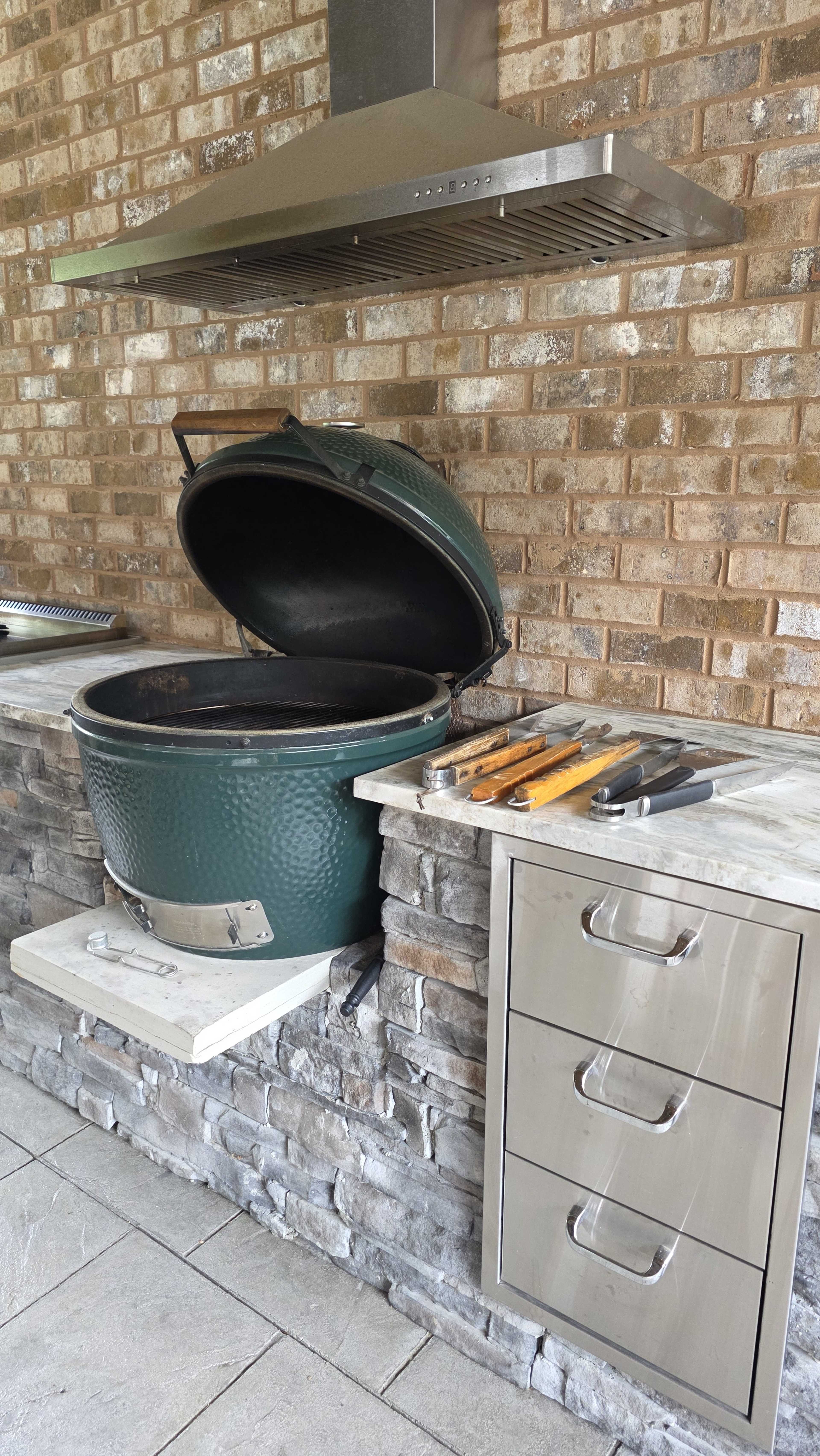 The image shows a green ceramic grill with an open lid, placed on a stone countertop beside stainless steel drawers and various grilling tools.