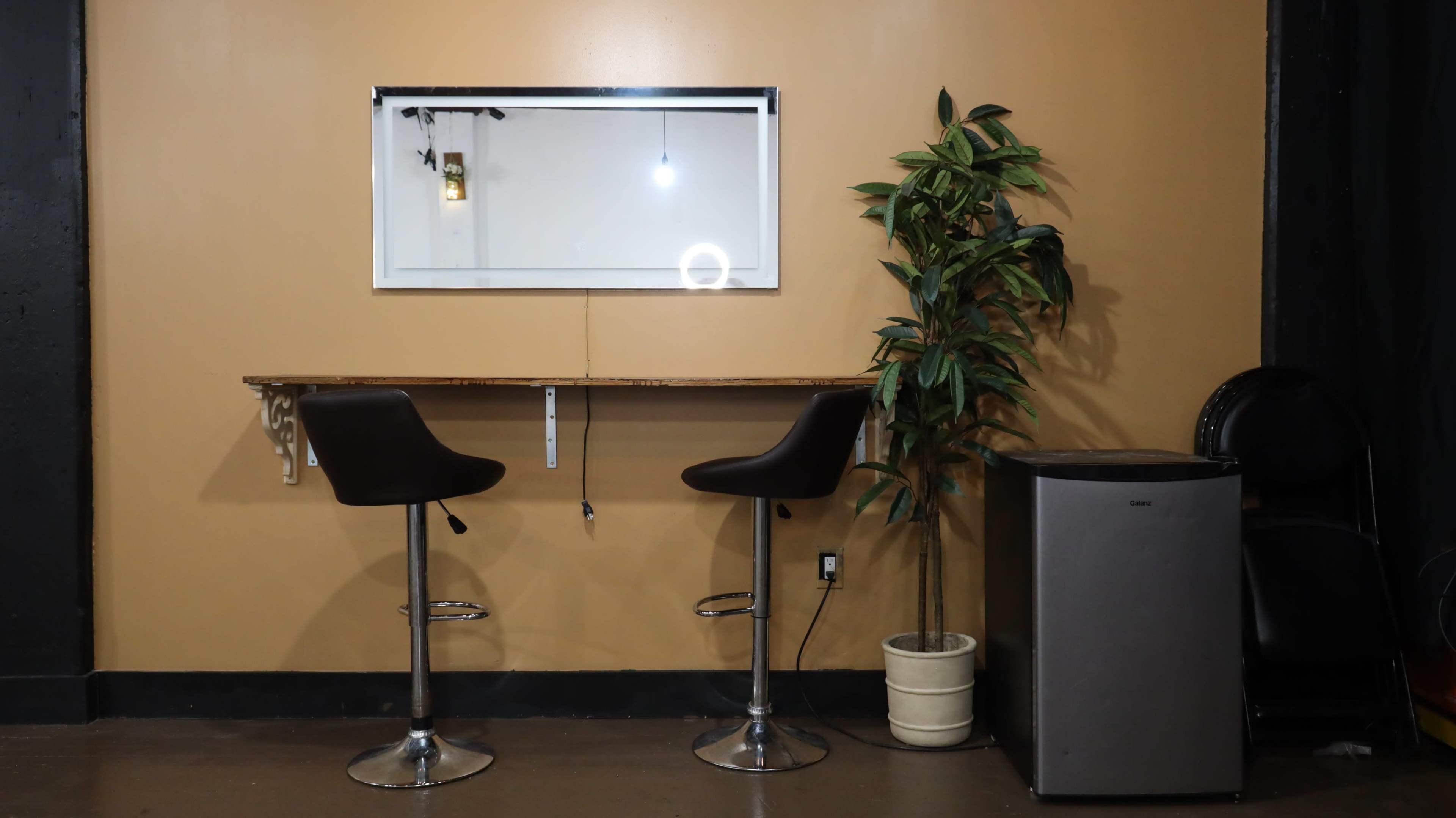 A wall with a wooden table and two black bar stools, a mirror above the table, a potted plant to the left, and a small gray fridge on the right.
