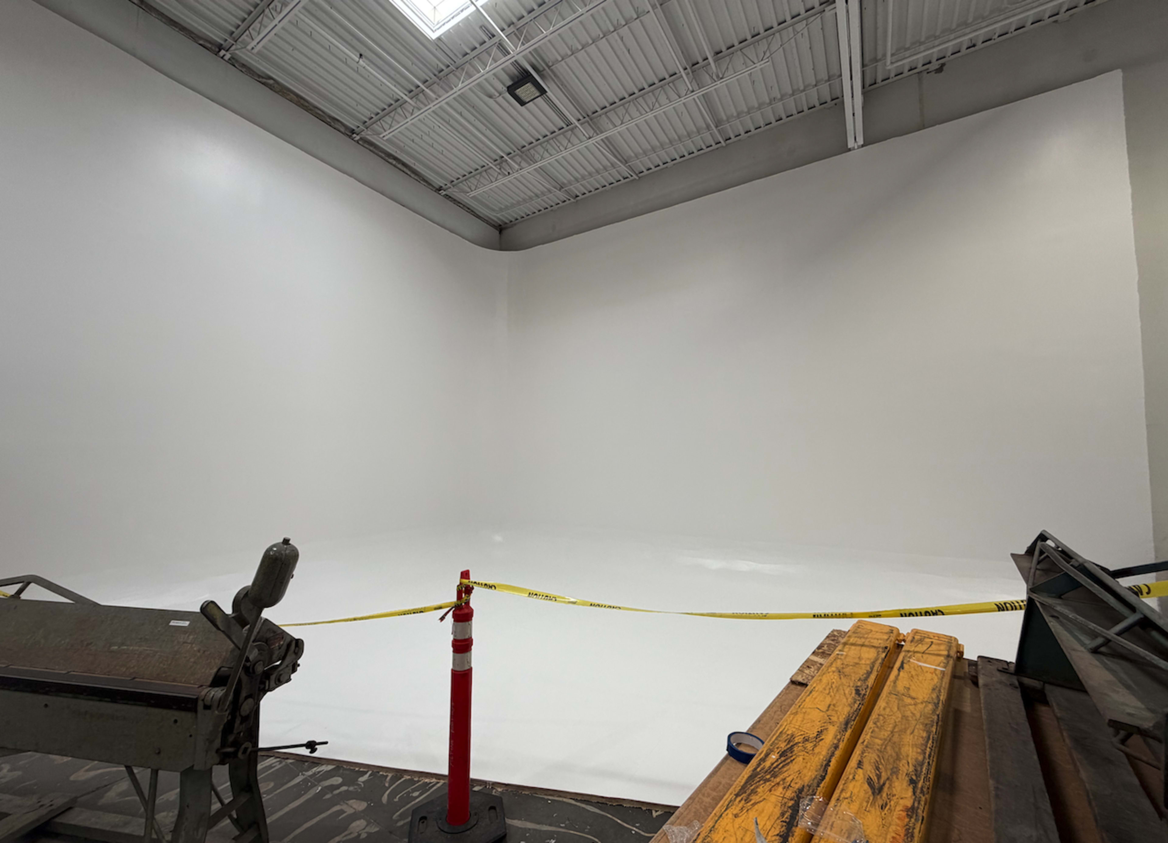 The image shows a large, empty white-walled room under construction, with caution tape and construction materials visible in the foreground.