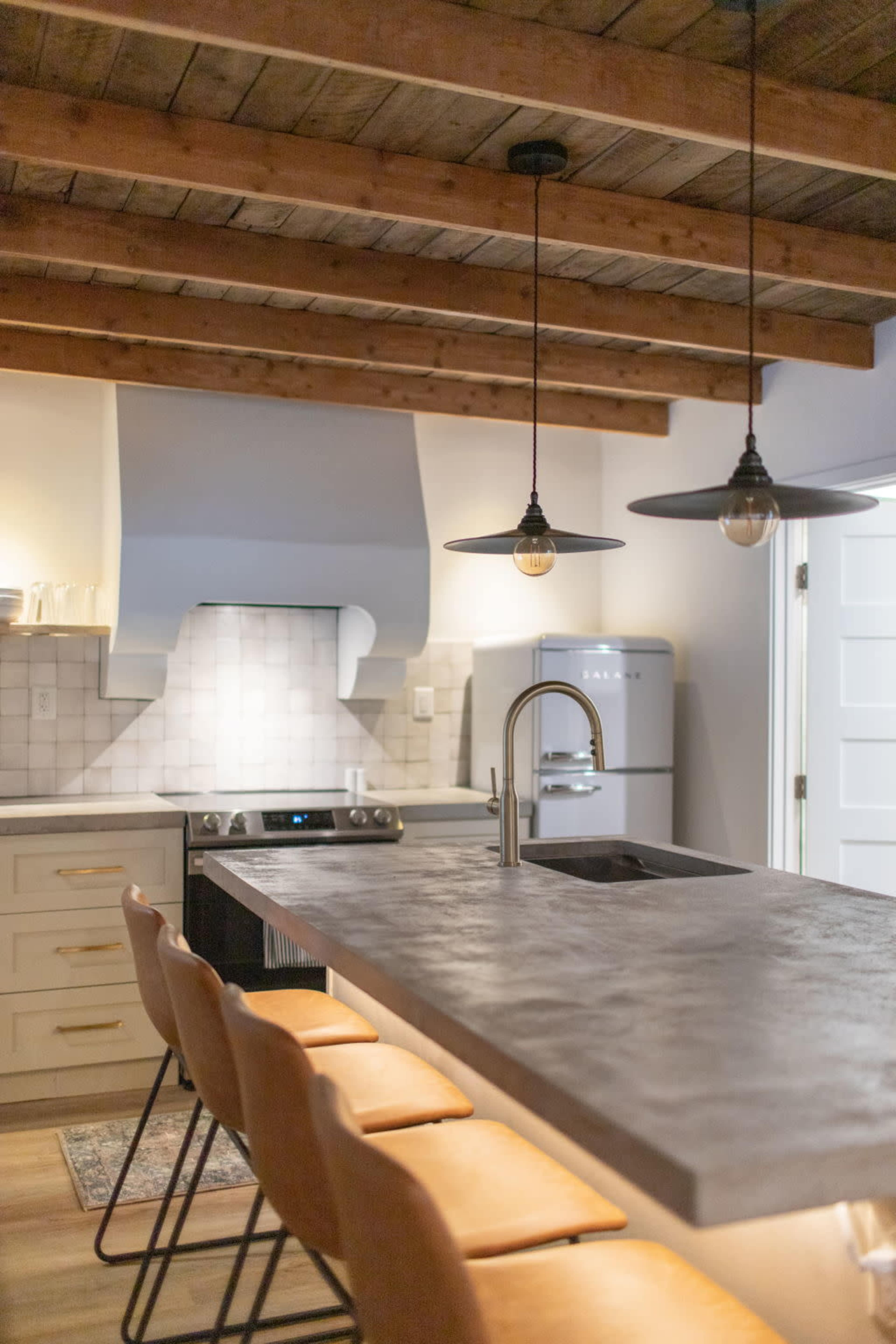 A modern kitchen features a large concrete island with bar stools, pendant lights, and wood beam ceilings.
