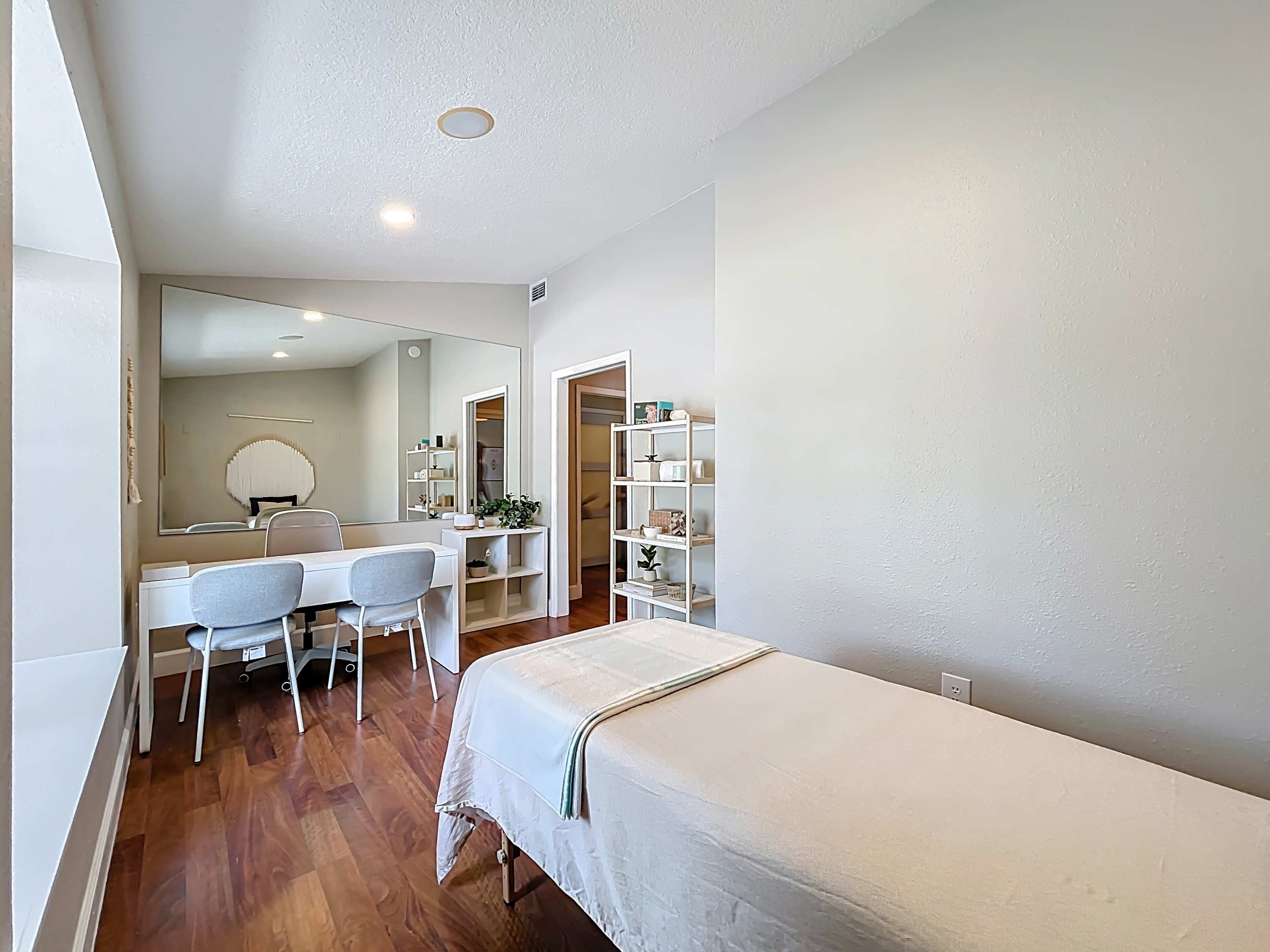 The image shows a serene treatment room featuring a massage table, two chairs, and a mirror along with minimalist shelving.