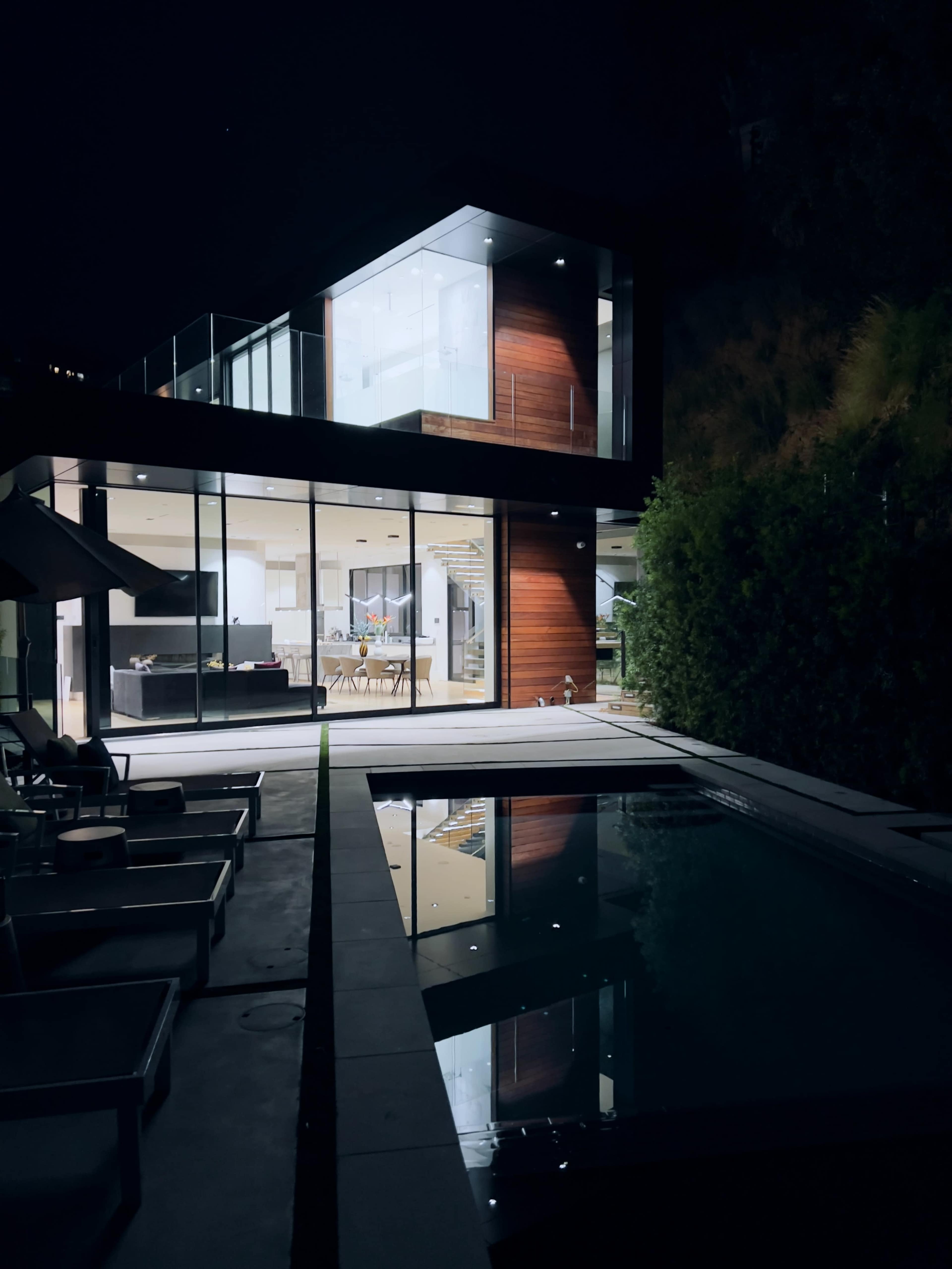 A modern house with large glass windows is illuminated at night, reflecting in a swimming pool surrounded by minimalist lounge chairs.