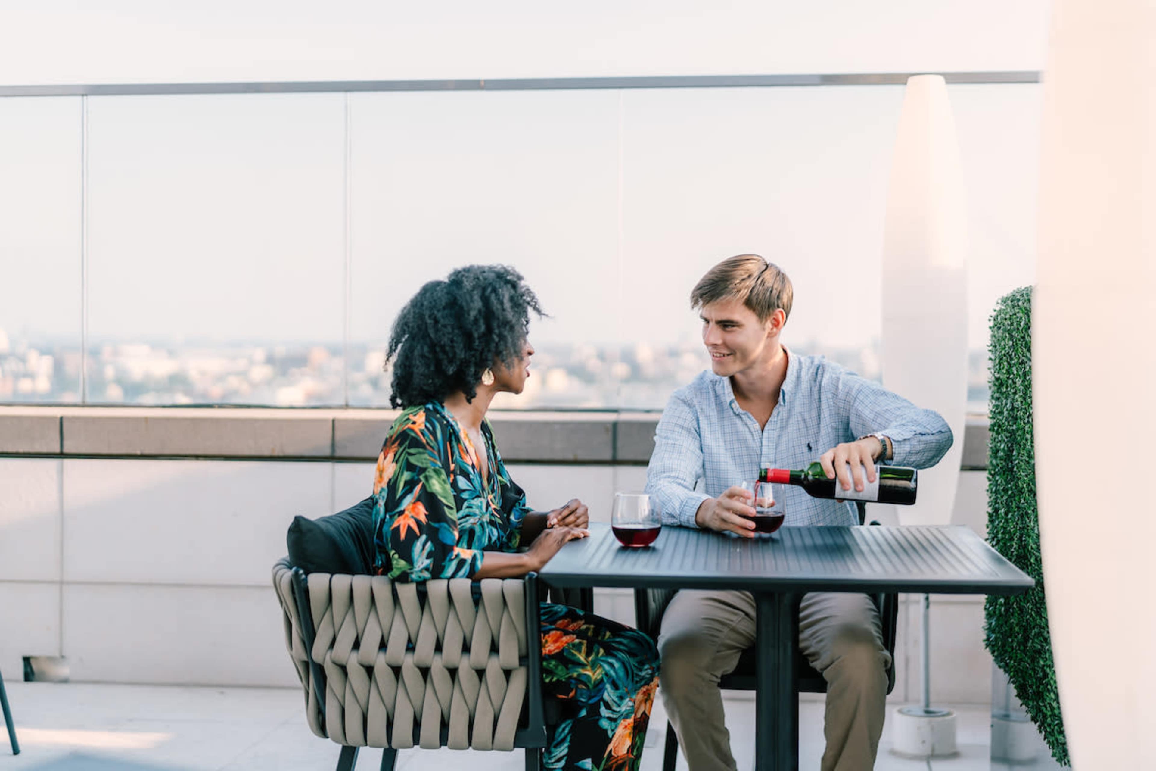 A man pours wine for a woman while they sit at a table on a rooftop overlooking a city skyline.