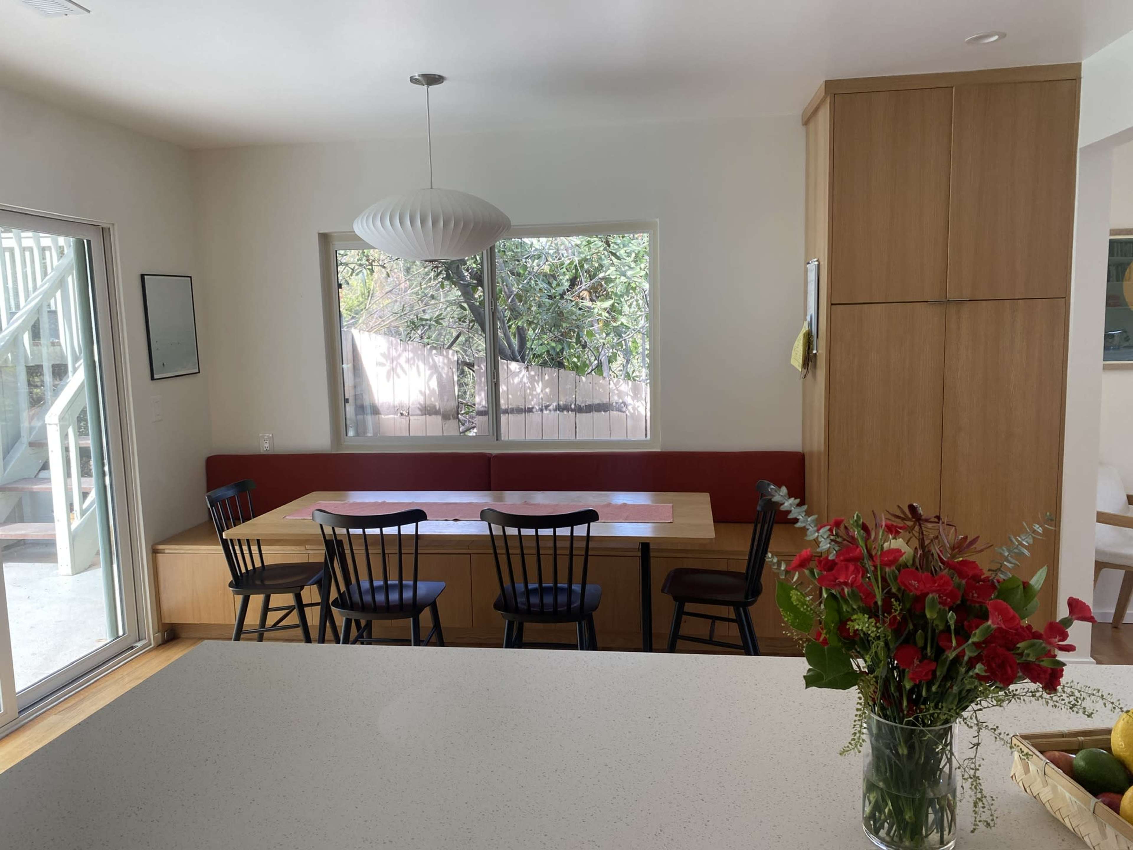 A modern kitchen features a dining area with a red bench, black chairs, and a large window overlooking greenery.