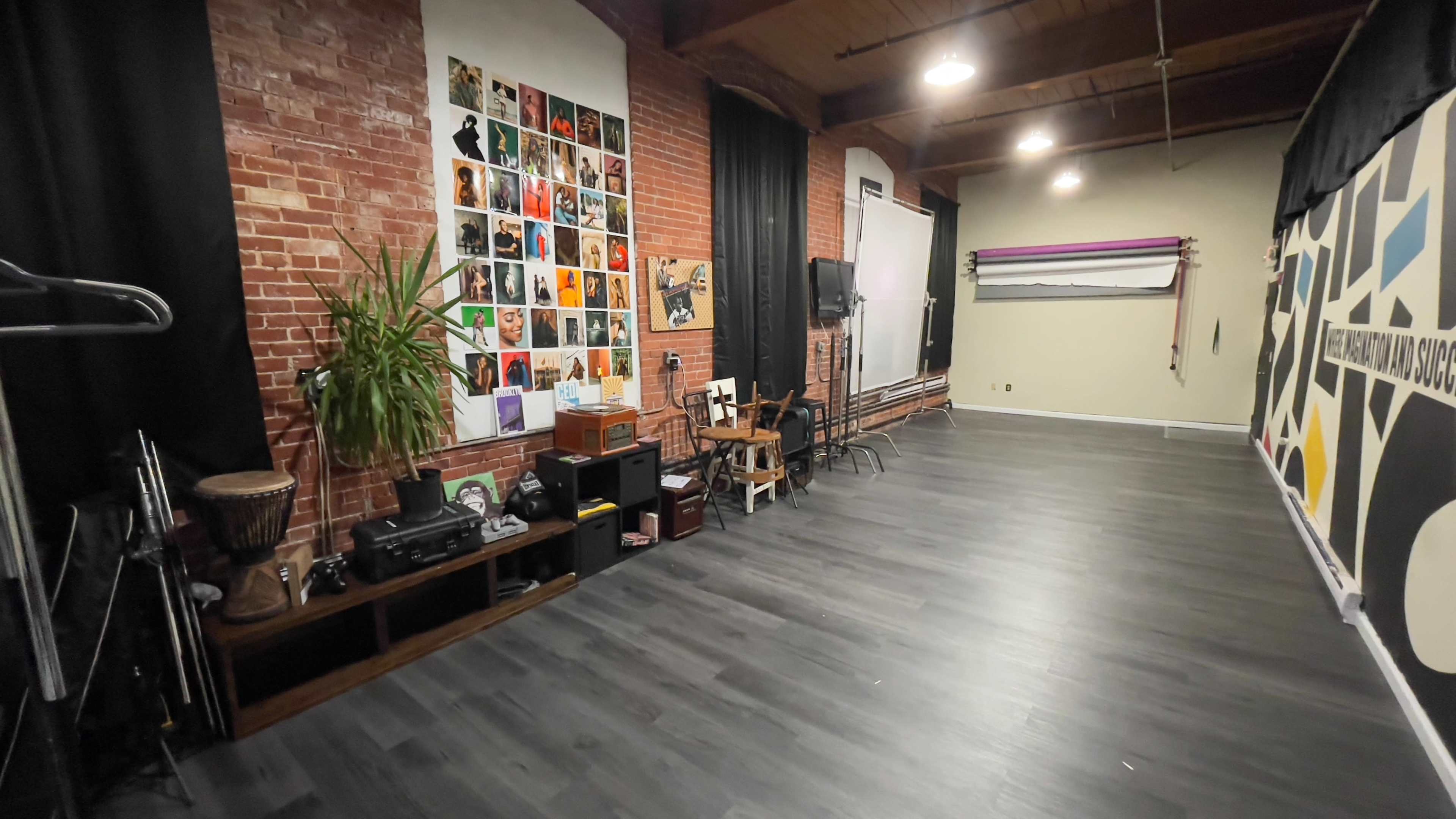 The image shows an art studio or rehearsal space with exposed brick walls, a variety of framed photos, a wooden floor, and various items including a plant, chairs, and a sound system.