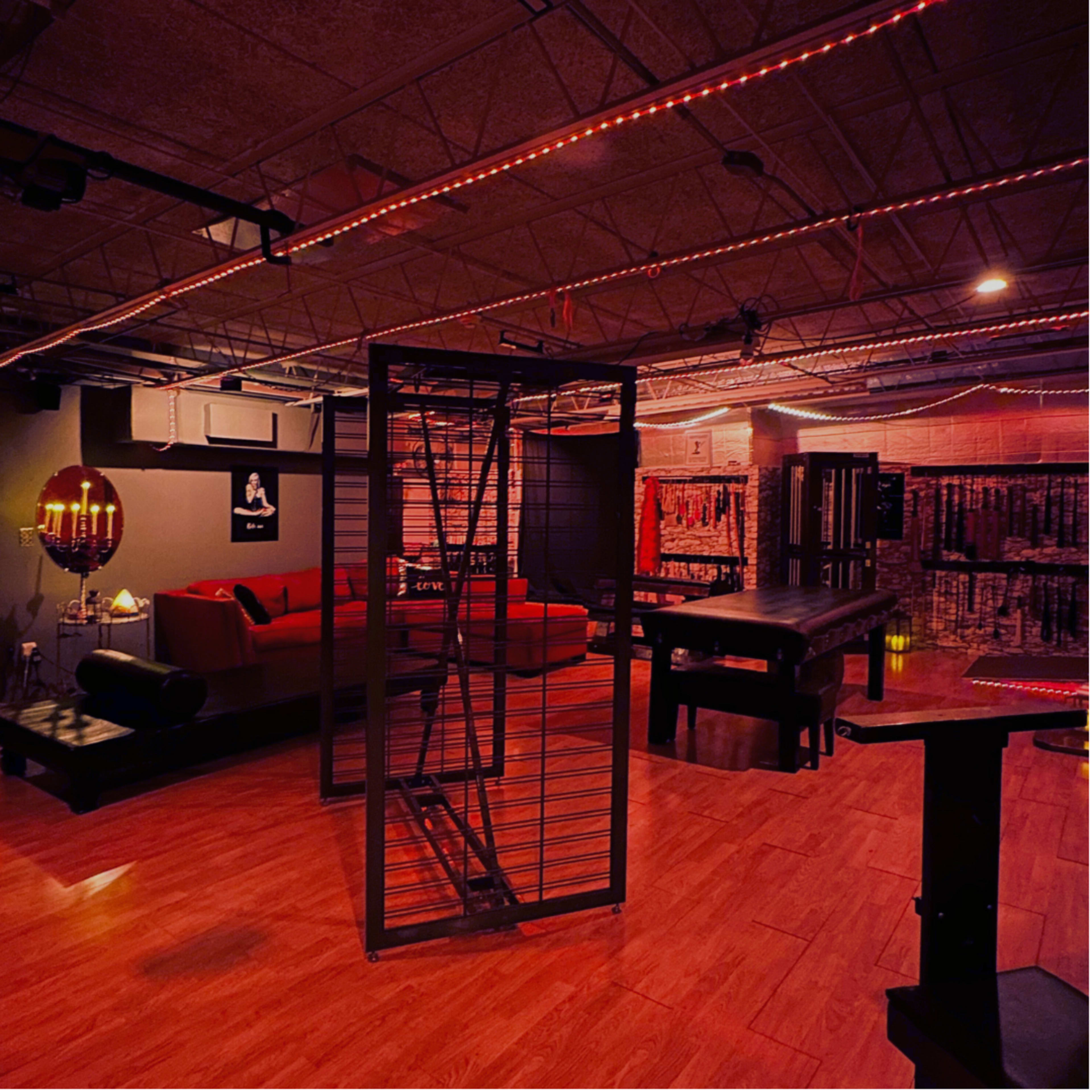 The image shows a dimly lit room with wooden flooring, featuring a red couch, a black table, and metal equipment arranged around the space.