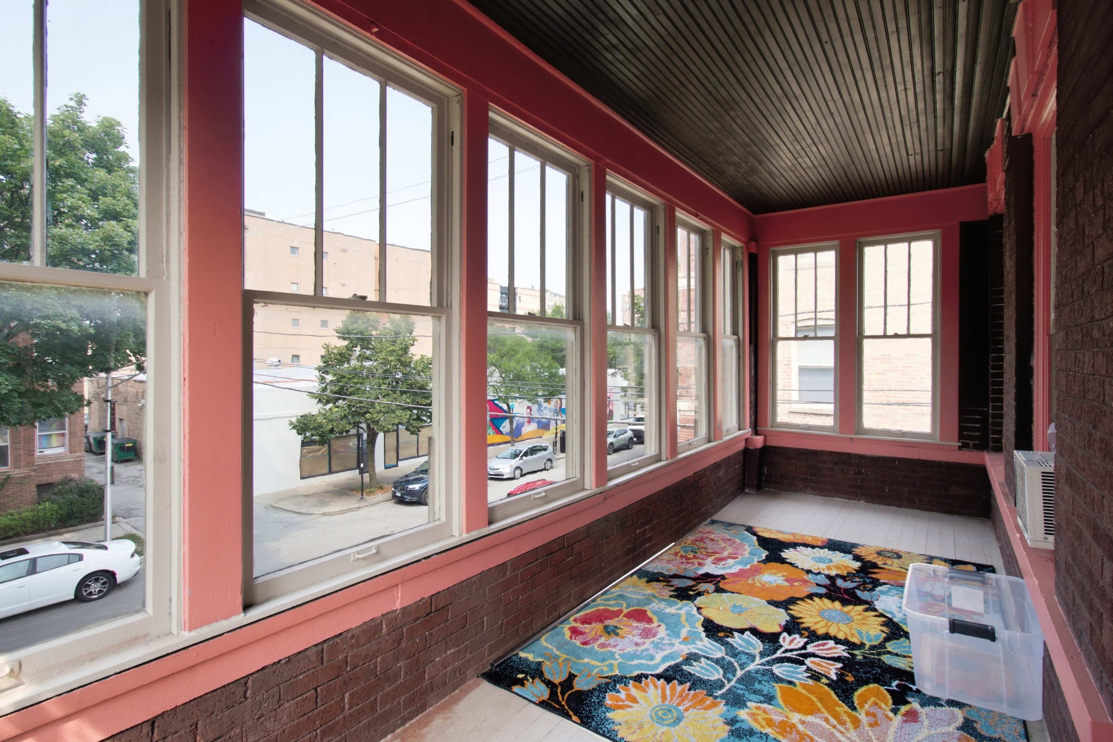 The image shows a brightly colored porch with large windows, a patterned rug on the floor, and a view of an outdoor street scene featuring trees and parked cars.