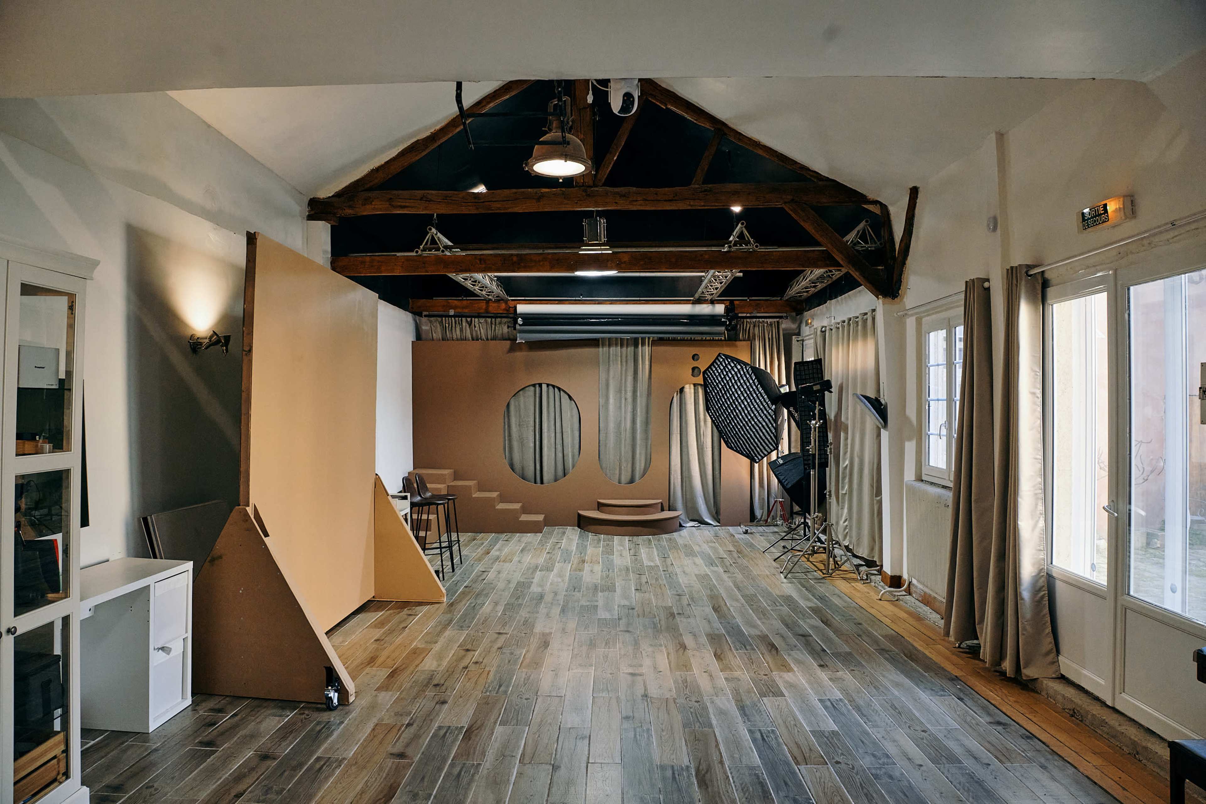 The image shows a spacious photography studio with wooden floors, exposed beams, and a setup that includes backdrops and lighting equipment.