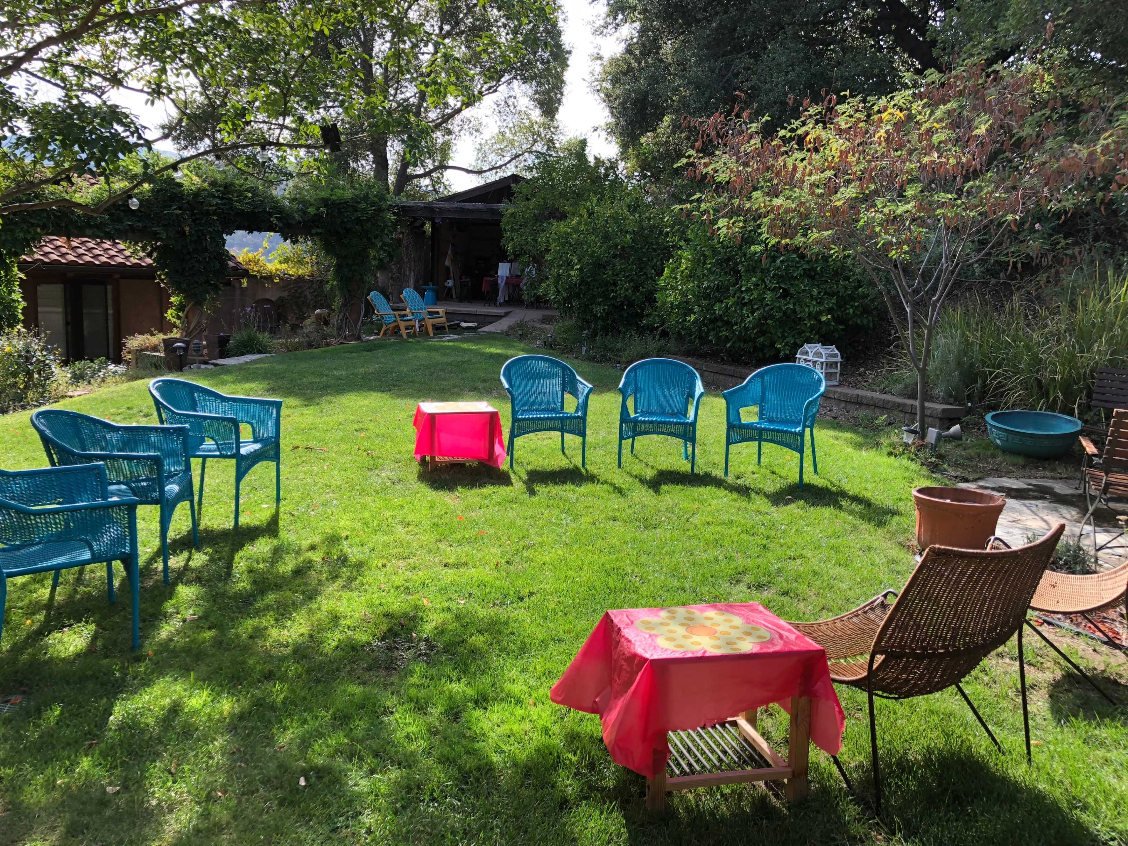 A backyard space features several blue chairs arranged in a circle around pink tables on green grass, with trees and a building in the background.