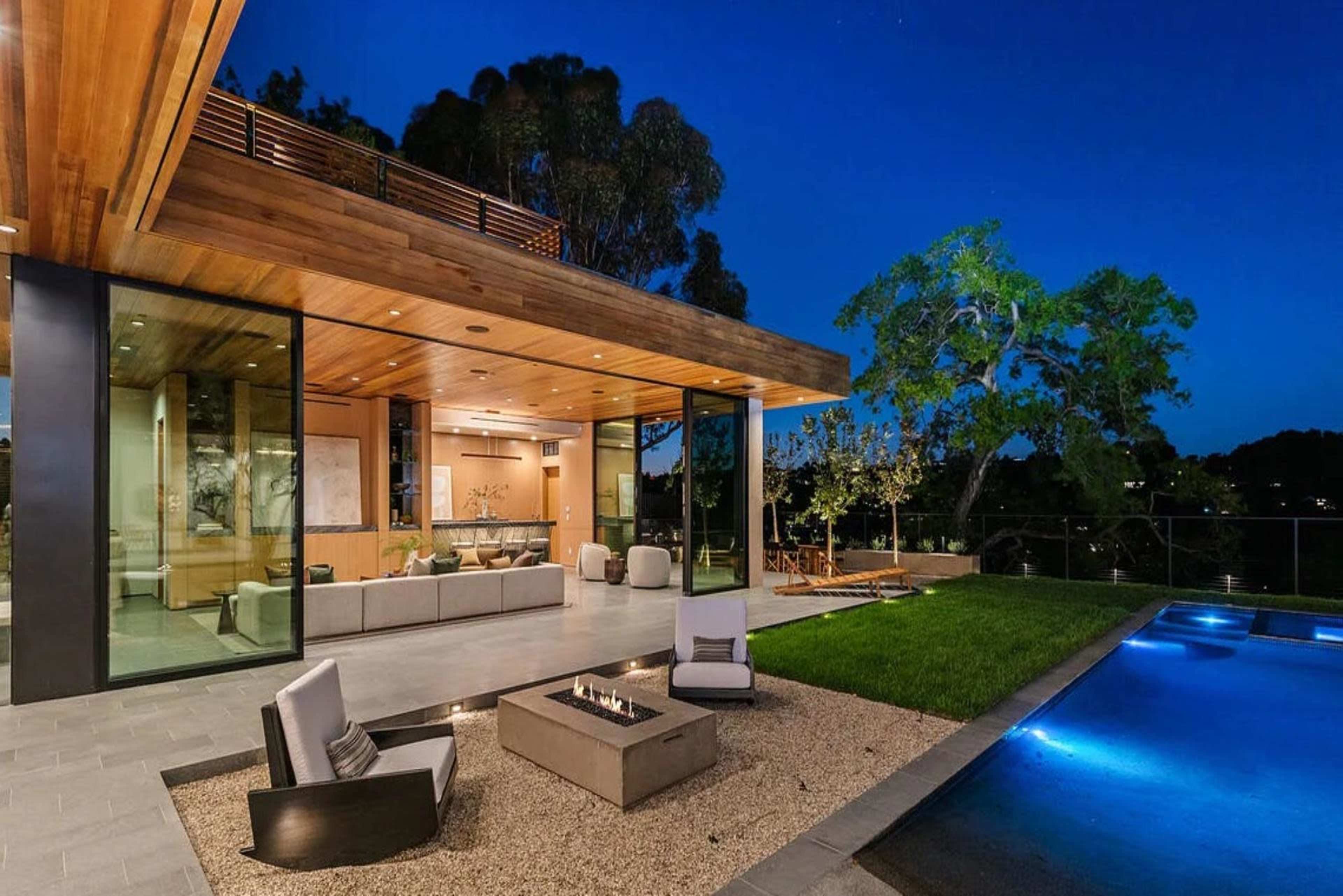 A modern house features a wooden overhang, large glass walls, and a pool with a fire pit in the outdoor living area.