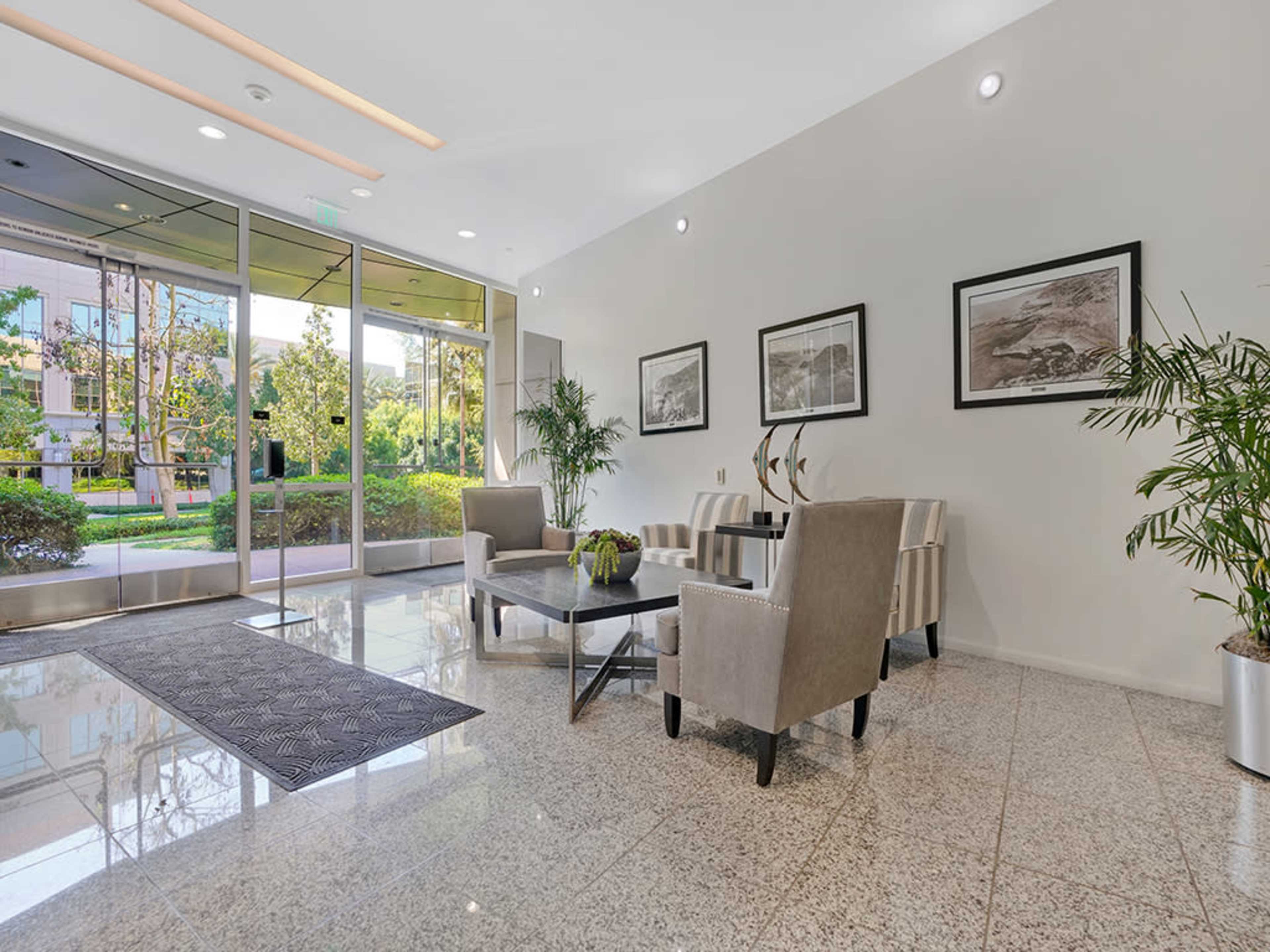 The image shows a modern lobby area with seating arrangements, a coffee table, and large windows revealing a garden view outside.