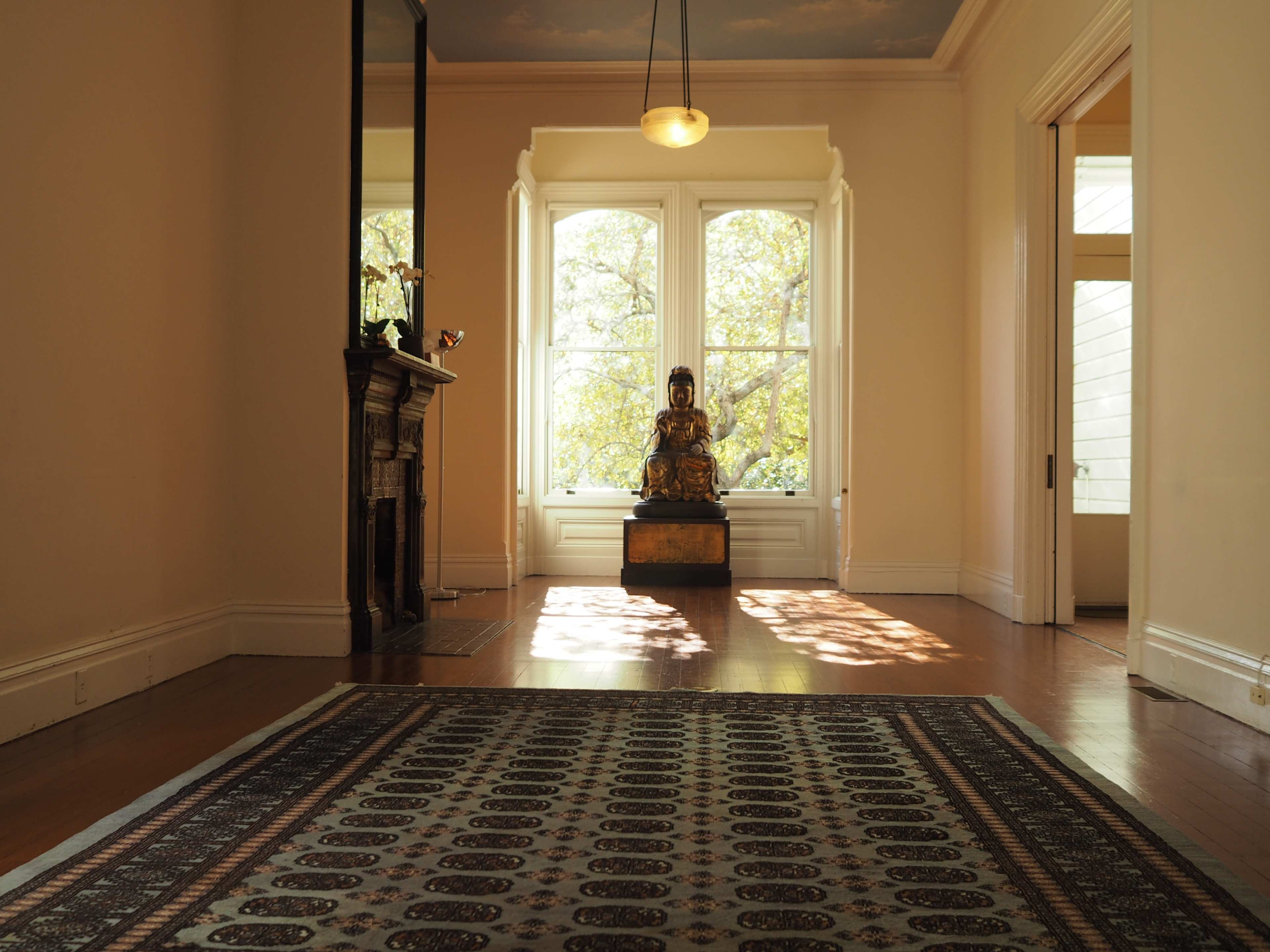 A spacious room features large windows that illuminate a seated statue in front of a decorative rug.