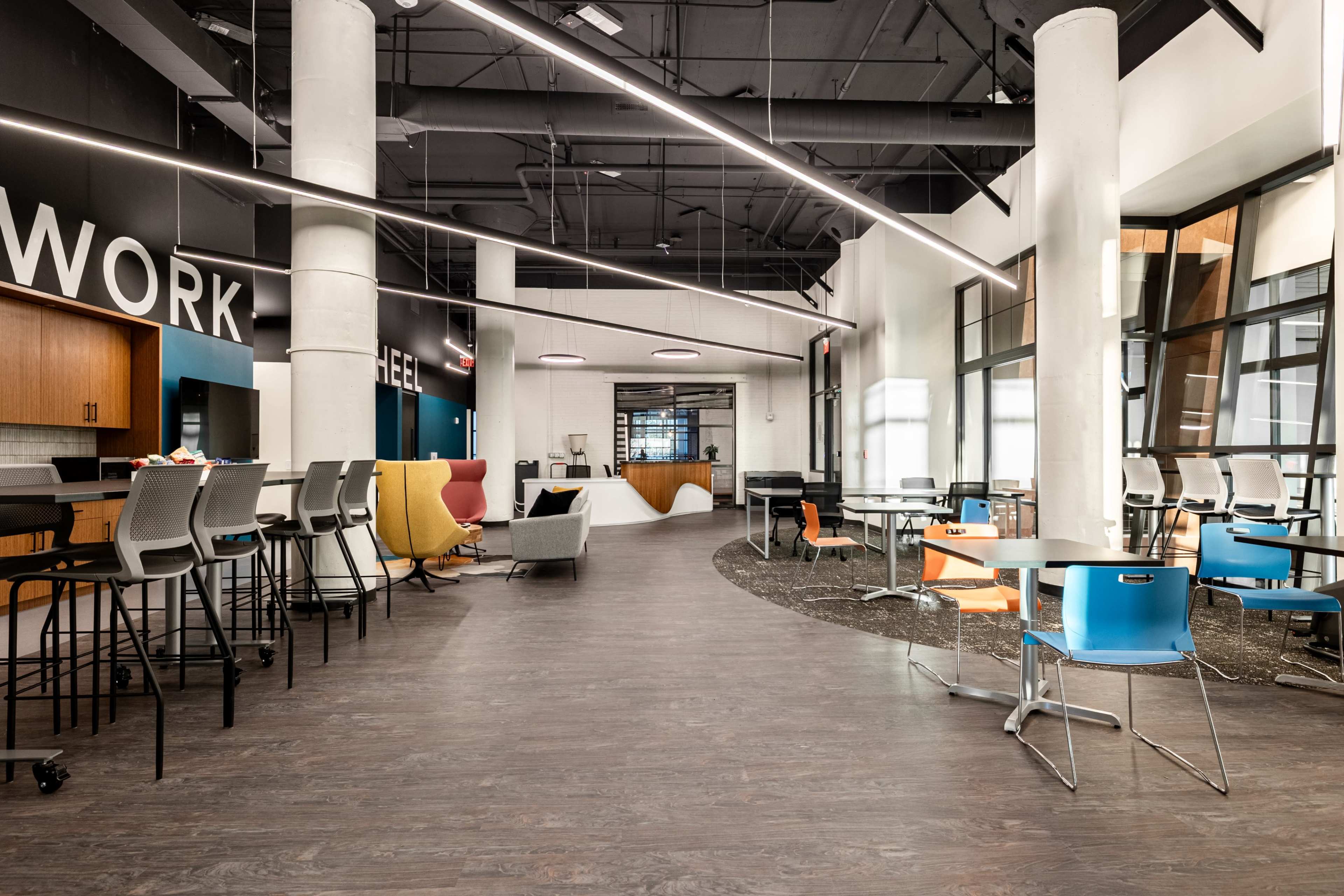The image shows a modern open workspace with various seating areas, including high tables and colorful chairs, illuminated by overhead lighting.