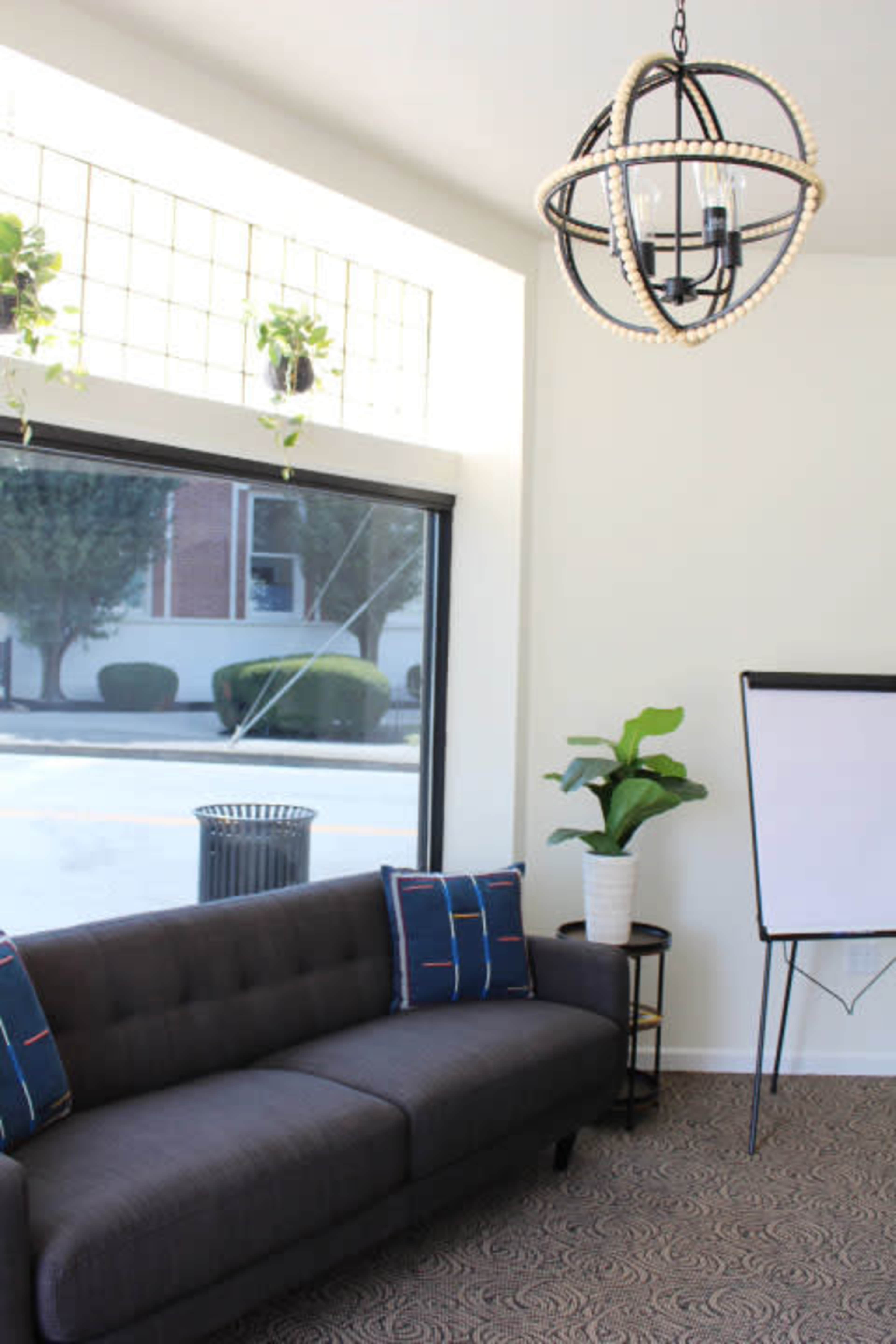 A modern seating area features a sofa with decorative pillows, a potted plant, a flip chart, and a large window that provides natural light.