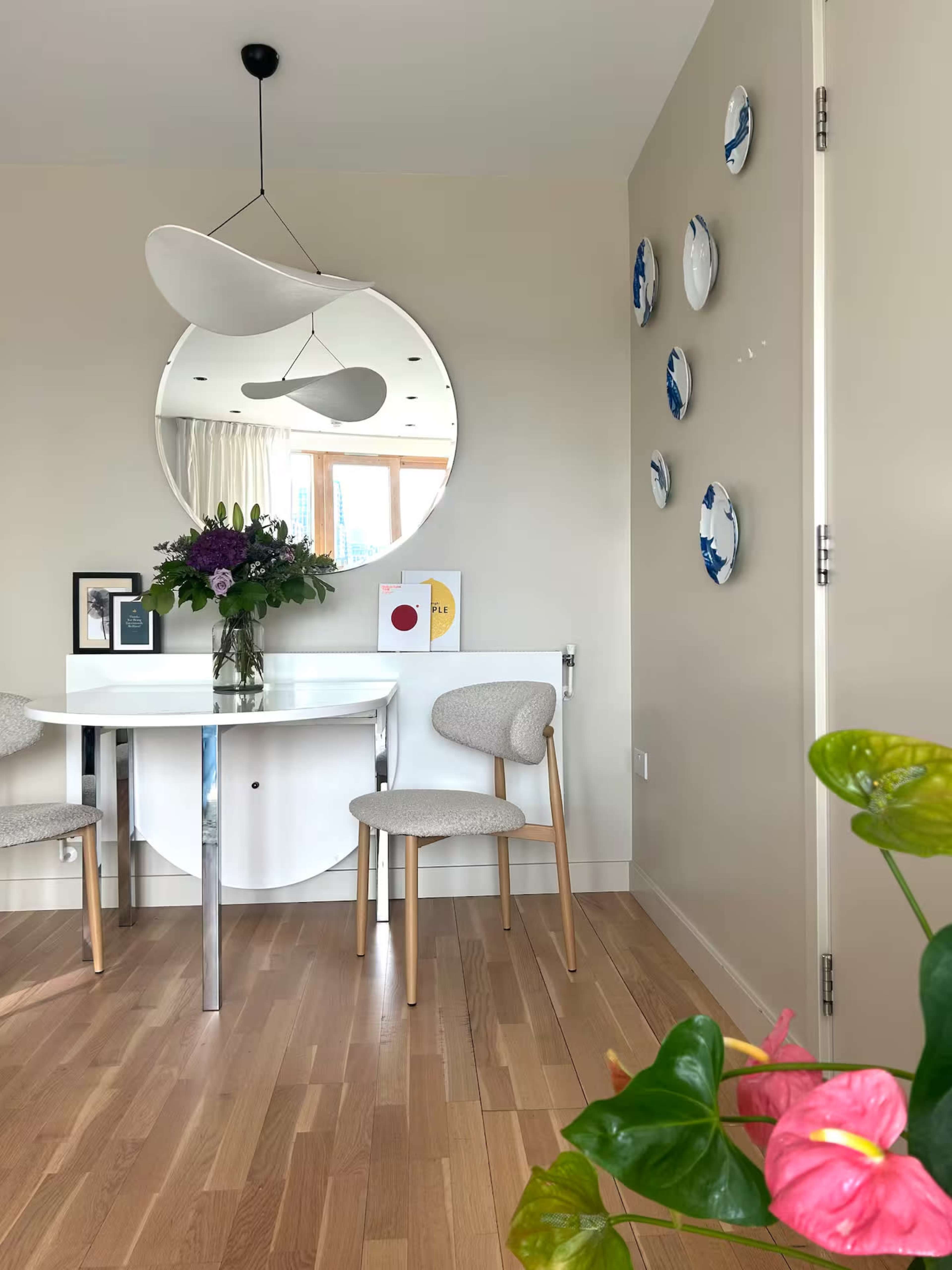 A modern dining area features a round mirror, a table with two chairs, floral decor, and decorative plates on the wall.