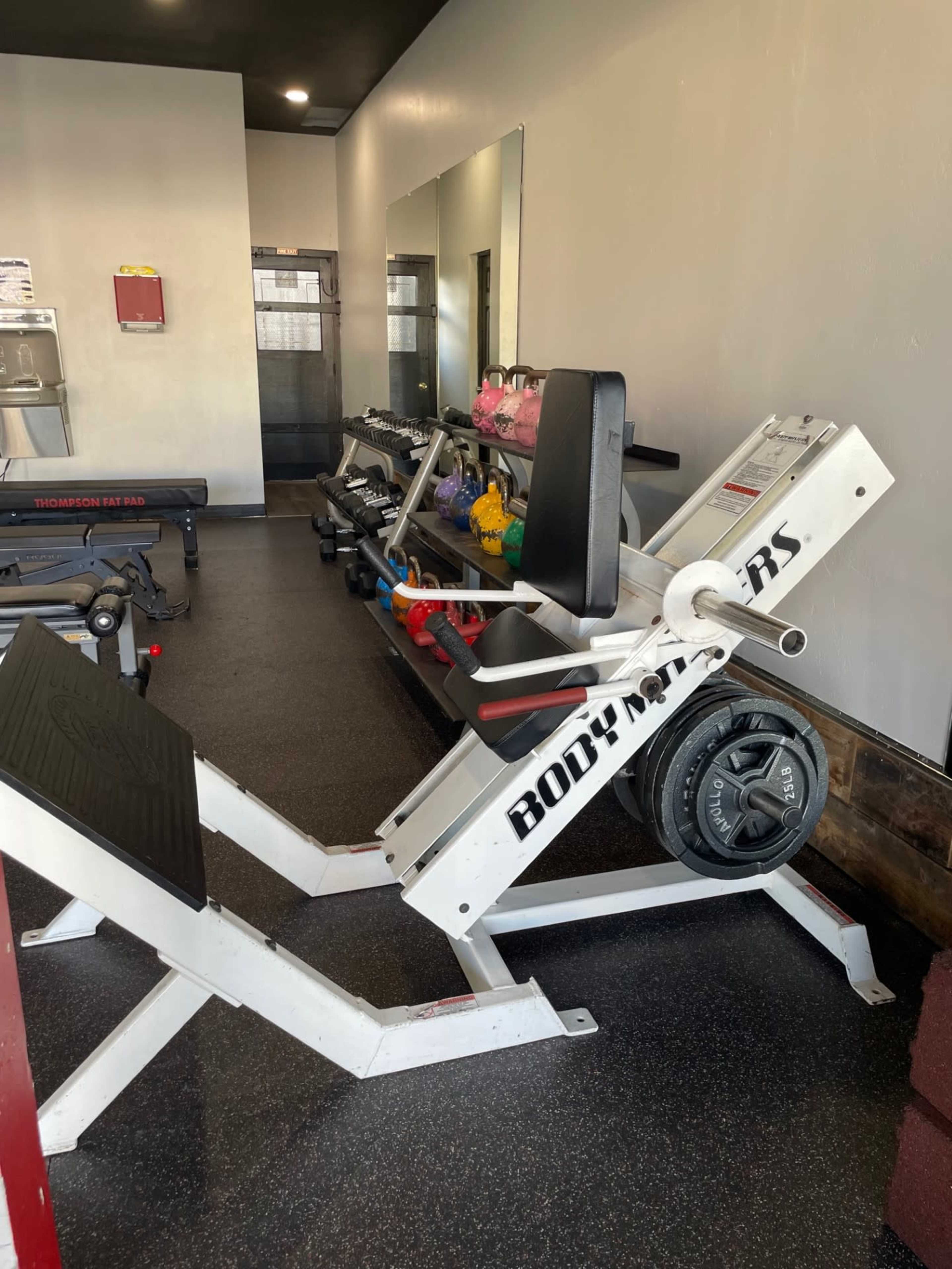 A gym interior featuring a leg press machine, free weights on a rack, and a mirrored wall.