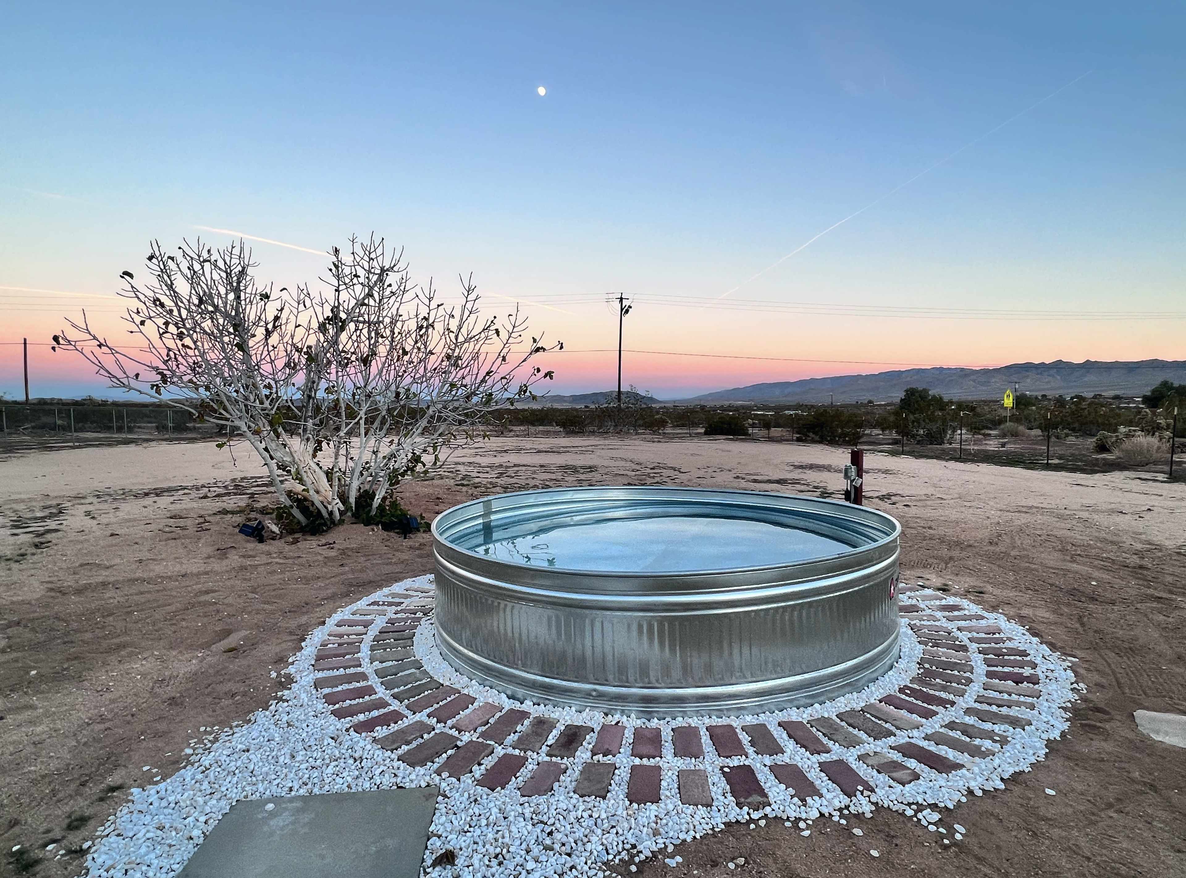 A circular metal water tank surrounded by a stone and gravel border sits in a barren landscape with a clear sky at dusk.