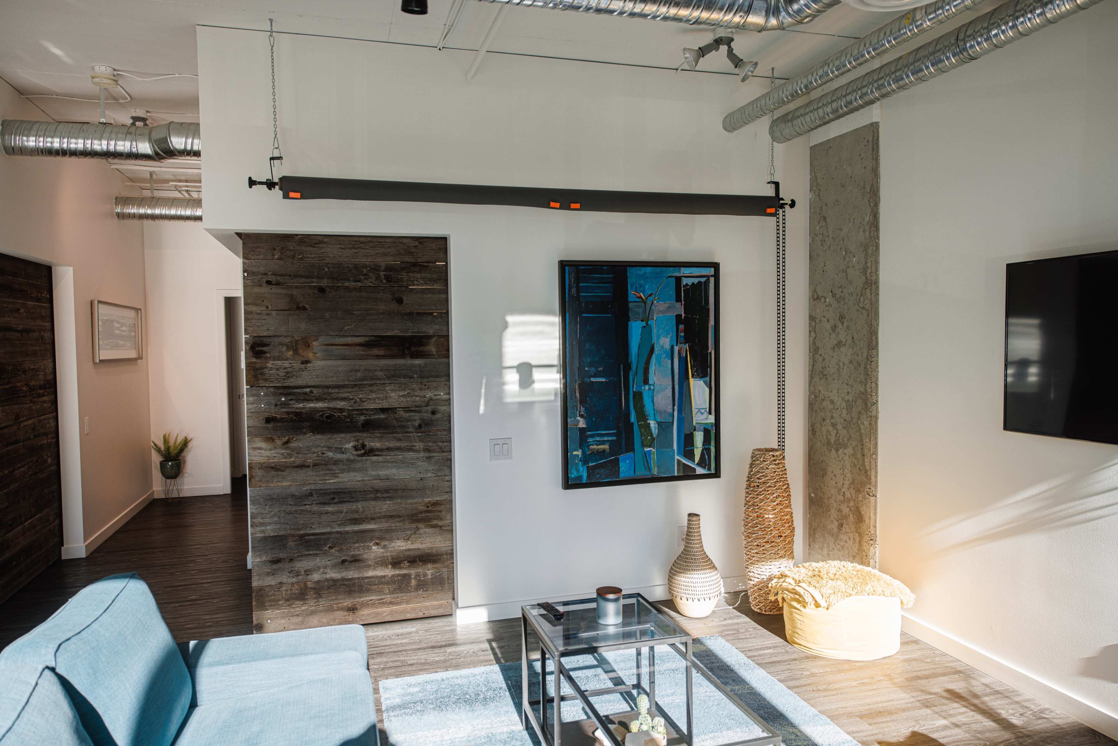 A modern interior space featuring a blue sofa, a glass coffee table, and a large framed artwork on the wall, with exposed ductwork and wooden accents.