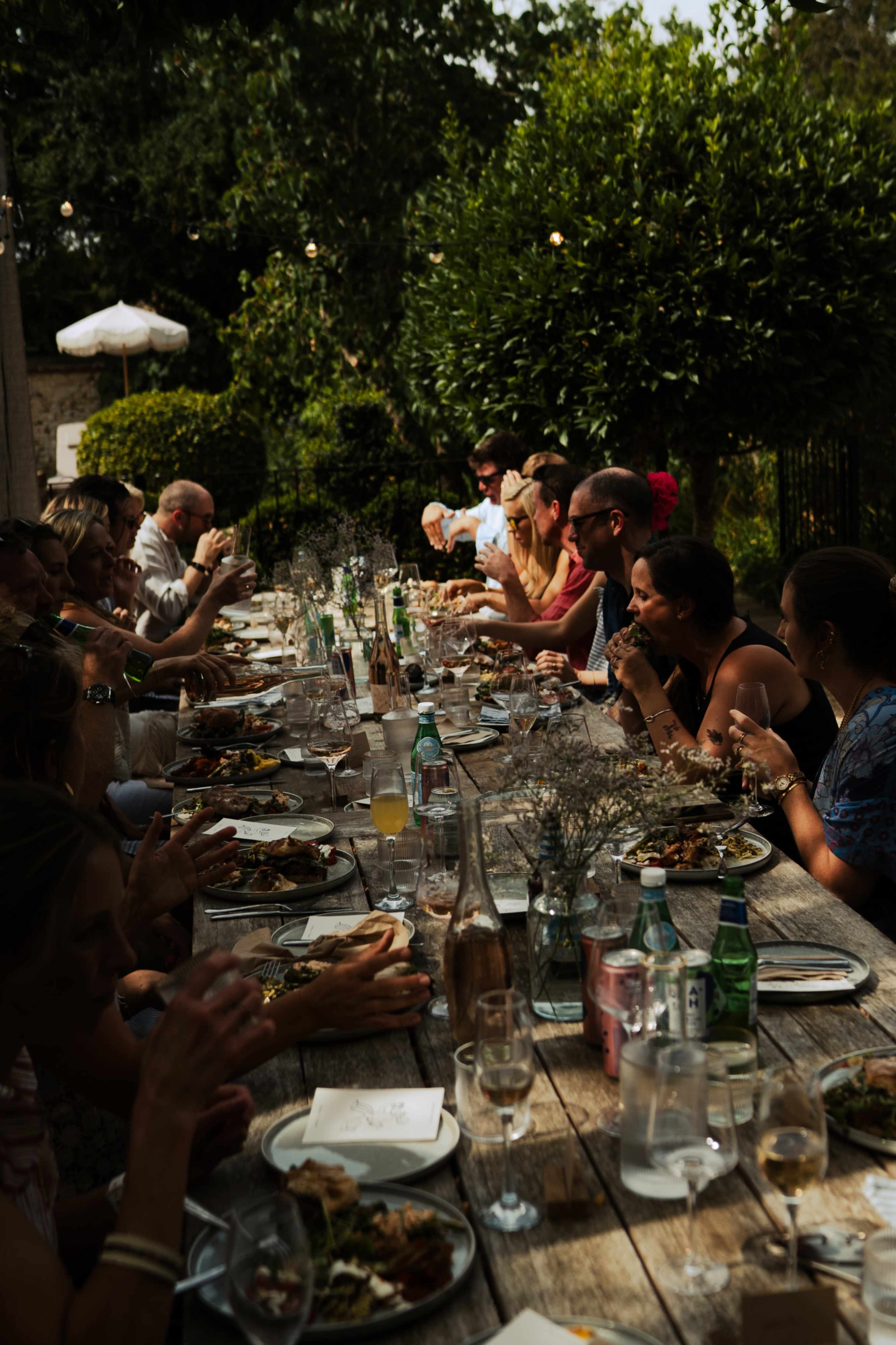A long dining table is filled with people enjoying a meal outdoors, surrounded by greenery and soft lighting.