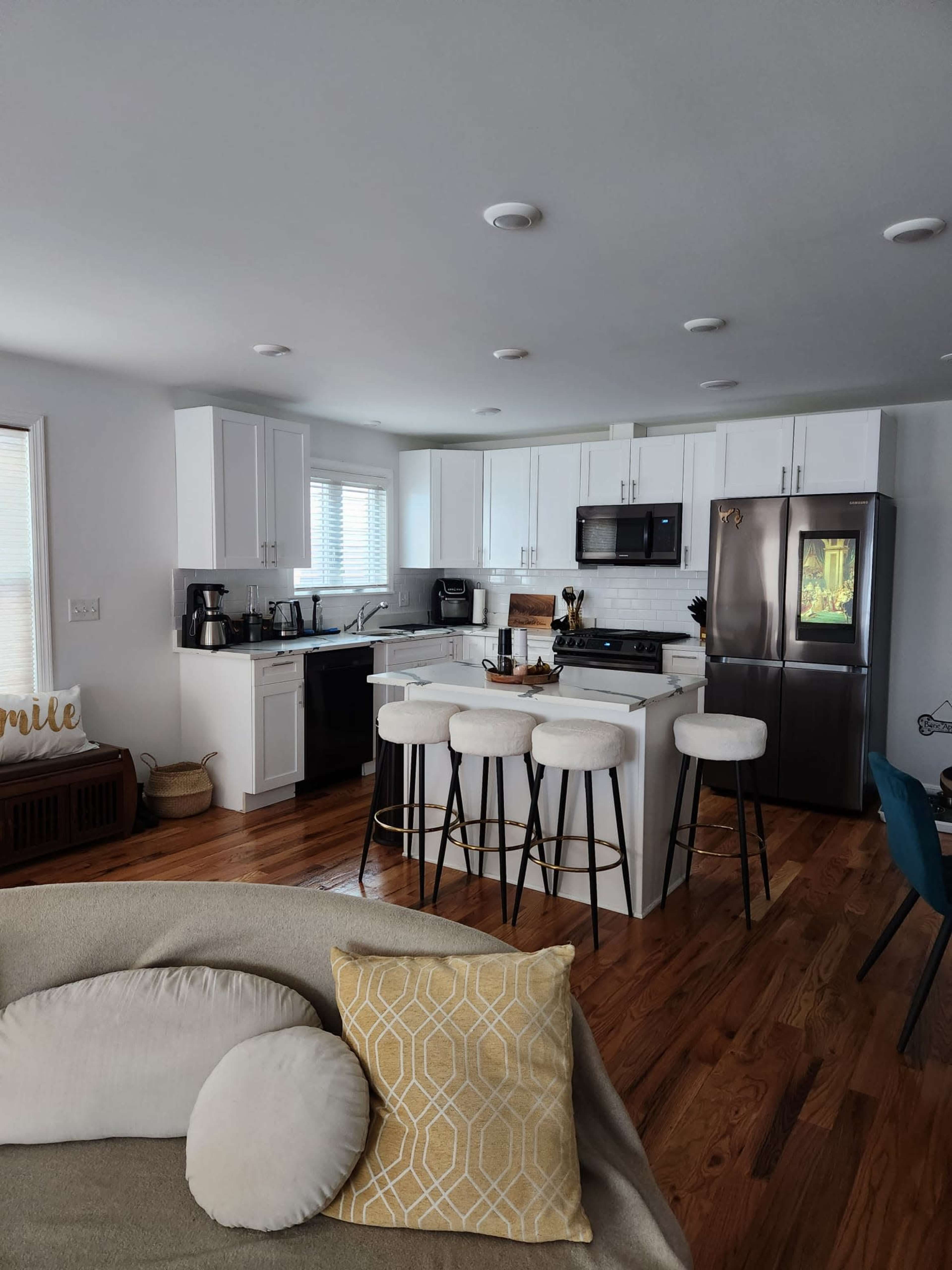 The image shows a modern kitchen with white cabinetry, a black refrigerator, and a breakfast bar with four stools, set against a backdrop of wooden flooring and large windows.