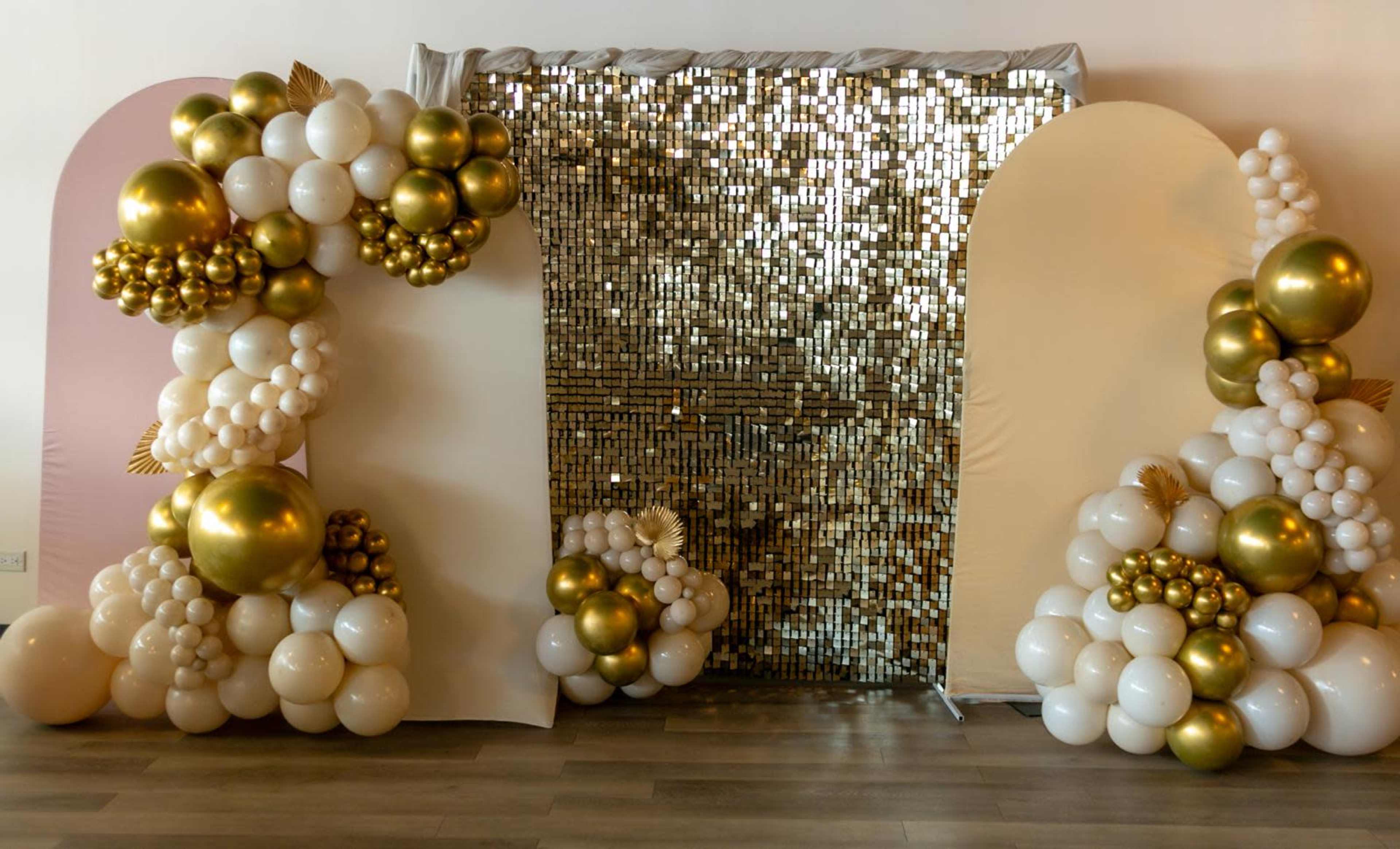 The image shows a decorative backdrop made of gold and white balloons arranged around a sparkling gold sequin wall.