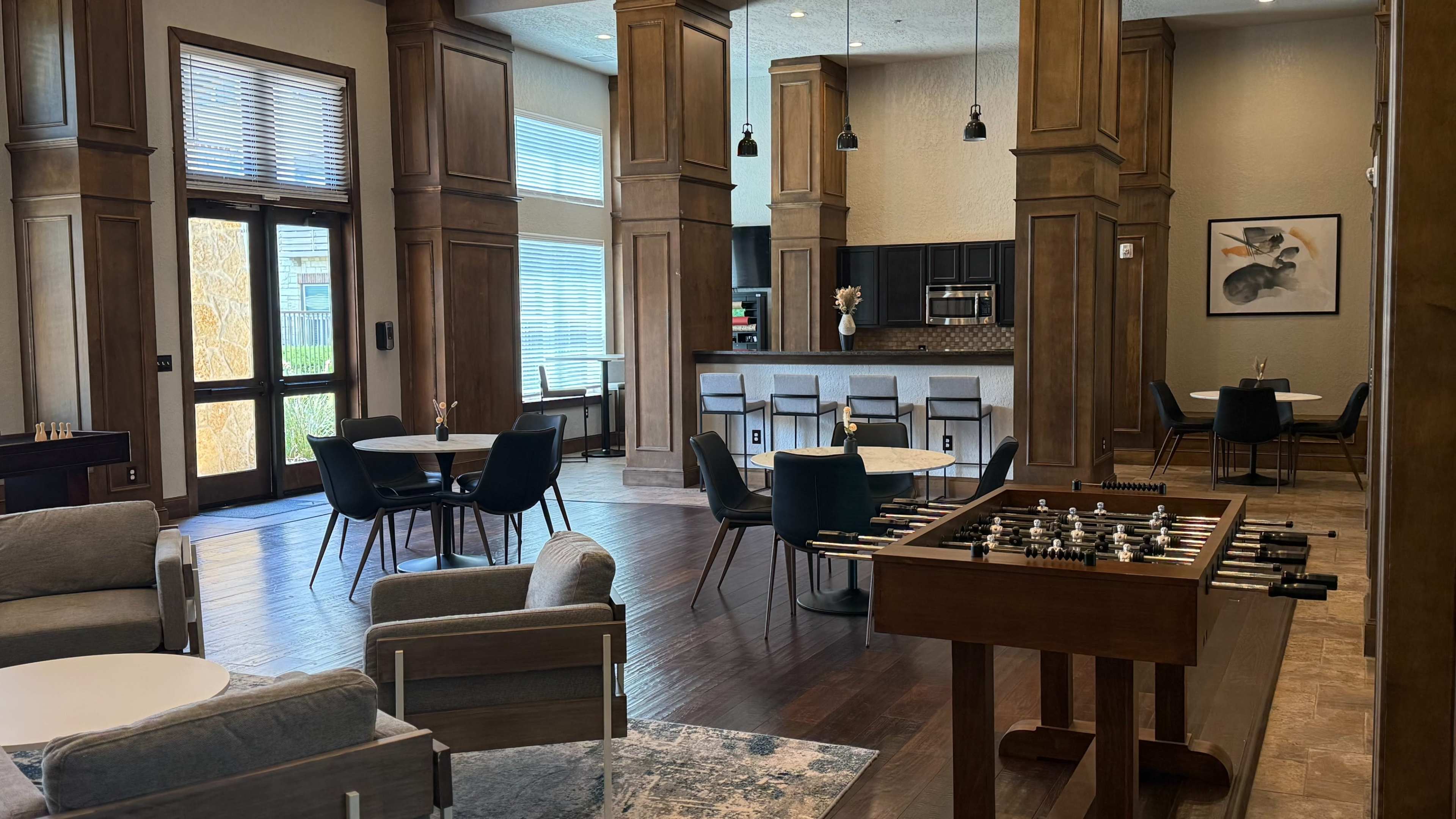 The image shows a modern lounge area with wooden pillars, seating arrangements, a foosball table, and a kitchenette in the background.