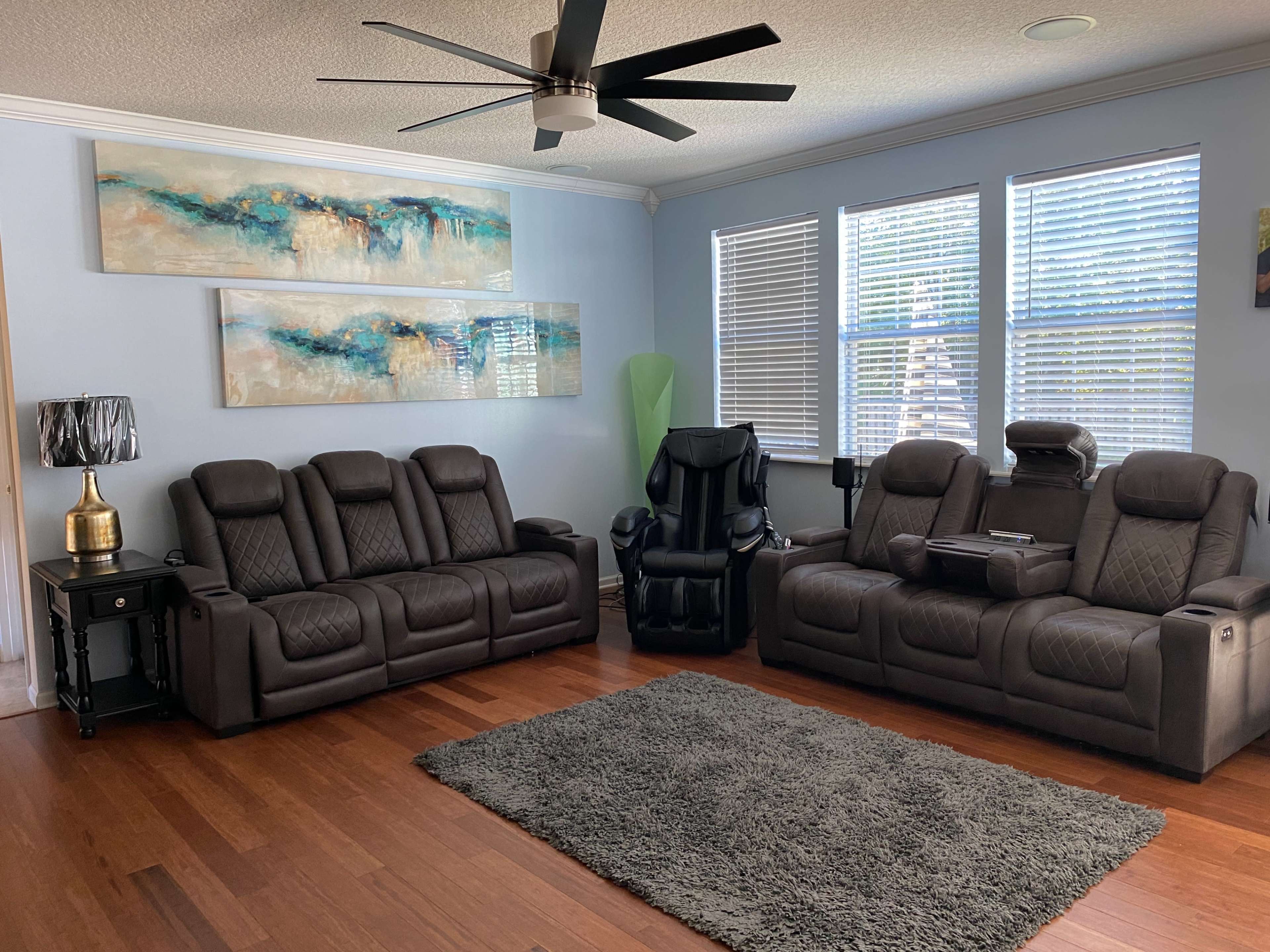 A living room with two black reclining sofas, a black gaming chair, and a large area rug on wooden flooring, complemented by wall art and bright, natural light coming through windows.