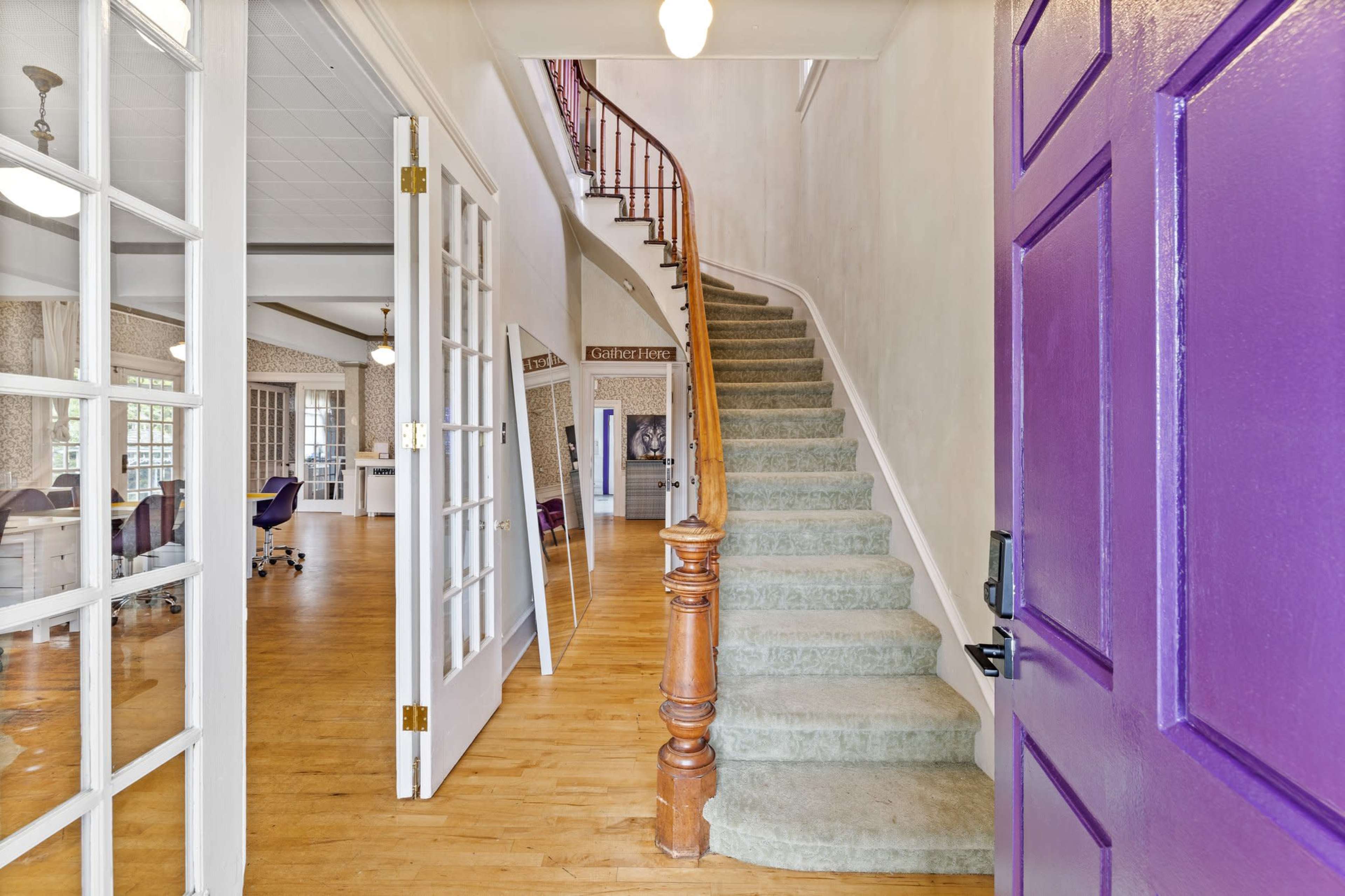 A well-lit entryway featuring a purple door, a curved staircase with wooden railing, and a spacious room visible through French doors.