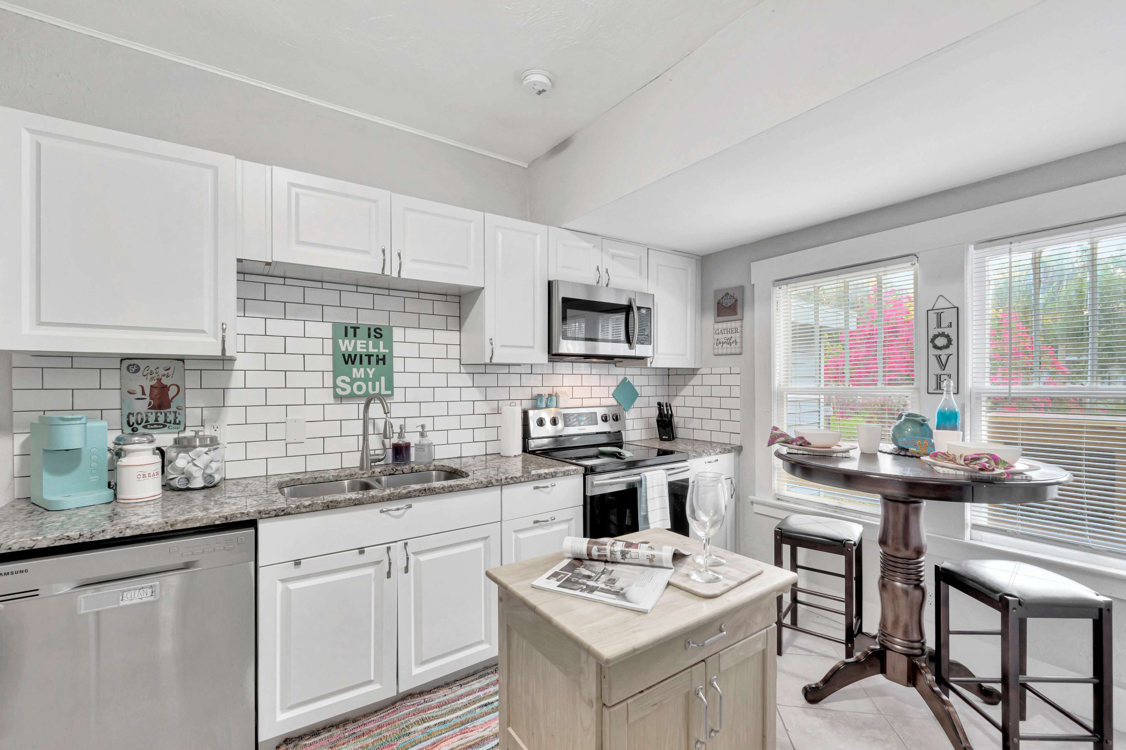 A modern kitchen with white cabinets, a gray countertop, a small dining table by the window, and tile backsplash.