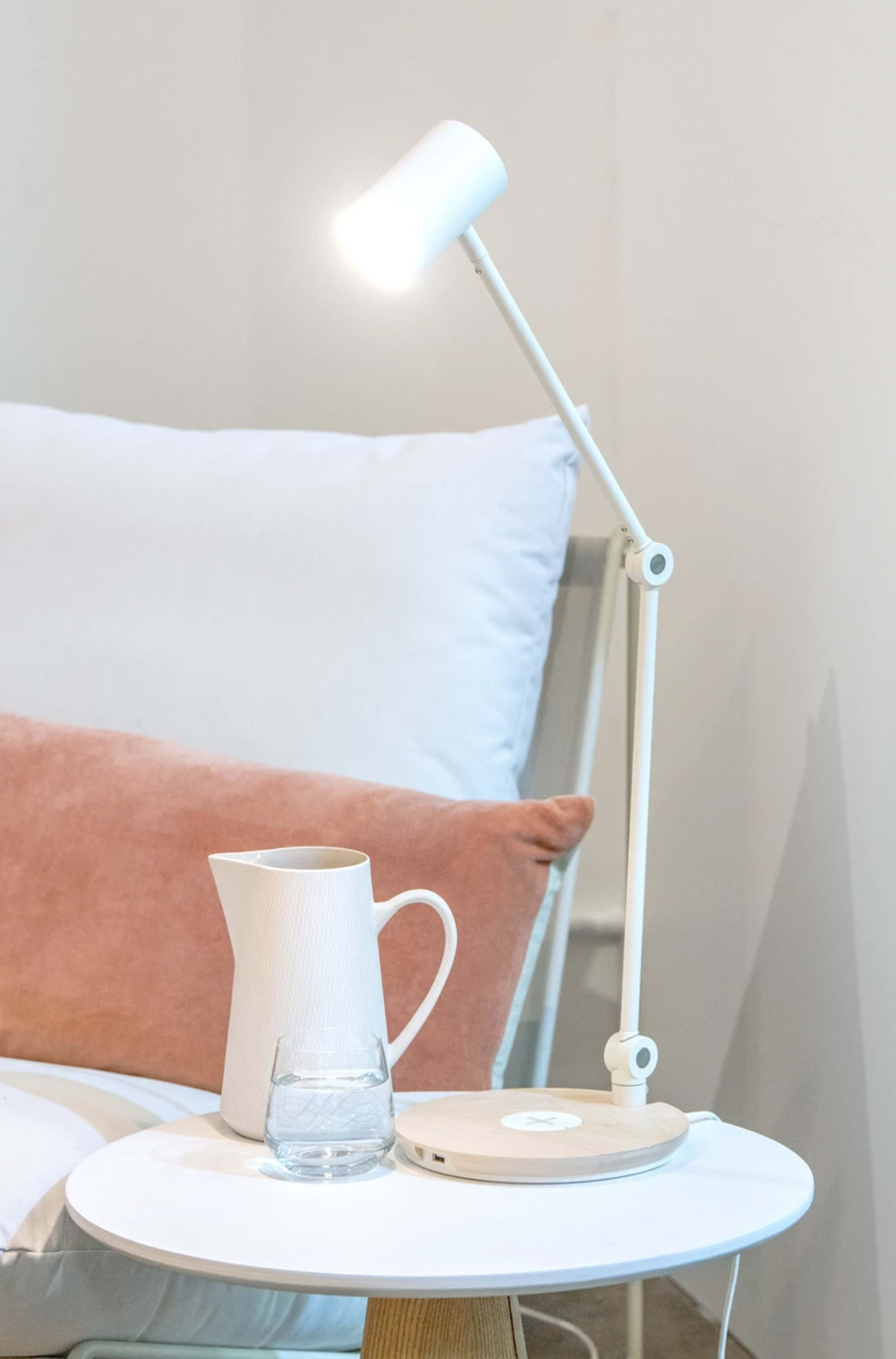 A white desk lamp illuminates a glass of water and a ceramic pitcher on a round side table beside a light-colored armchair with a pink cushion.