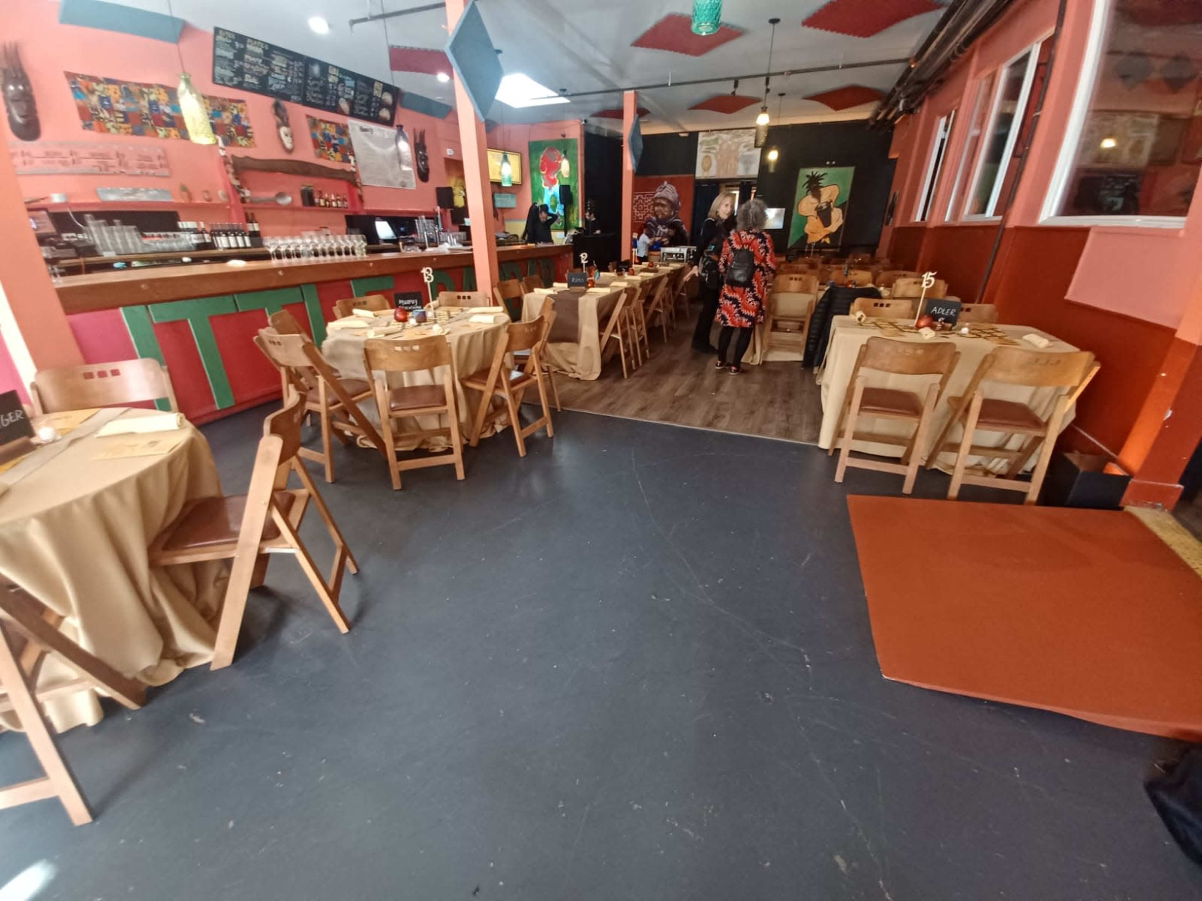 The interior of a restaurant with wooden tables and chairs set up, featuring a brightly colored decor and a bar area in the background.
